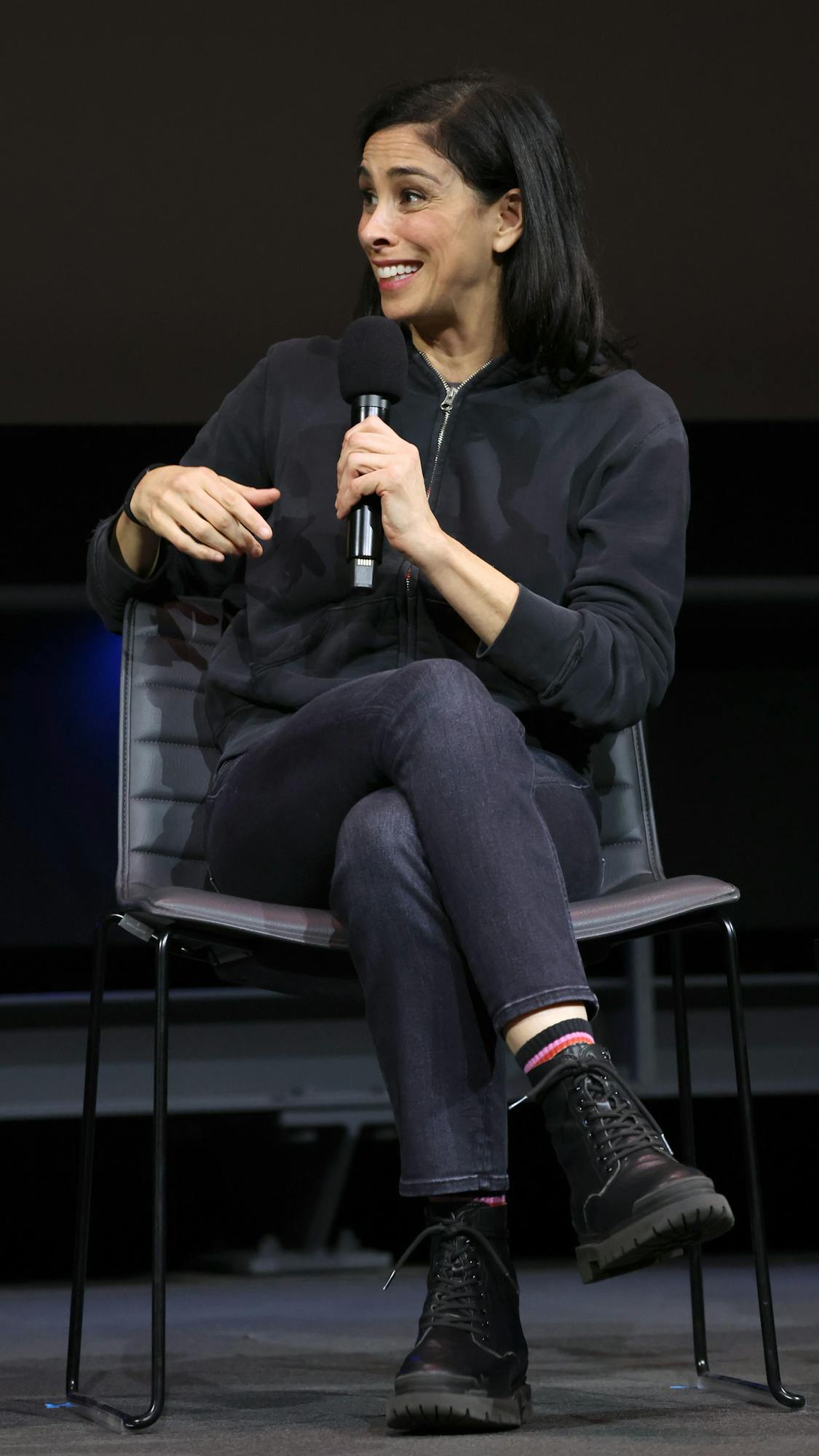 Sarah Silverman wears jeans and a black sweater and laughs into a mic.