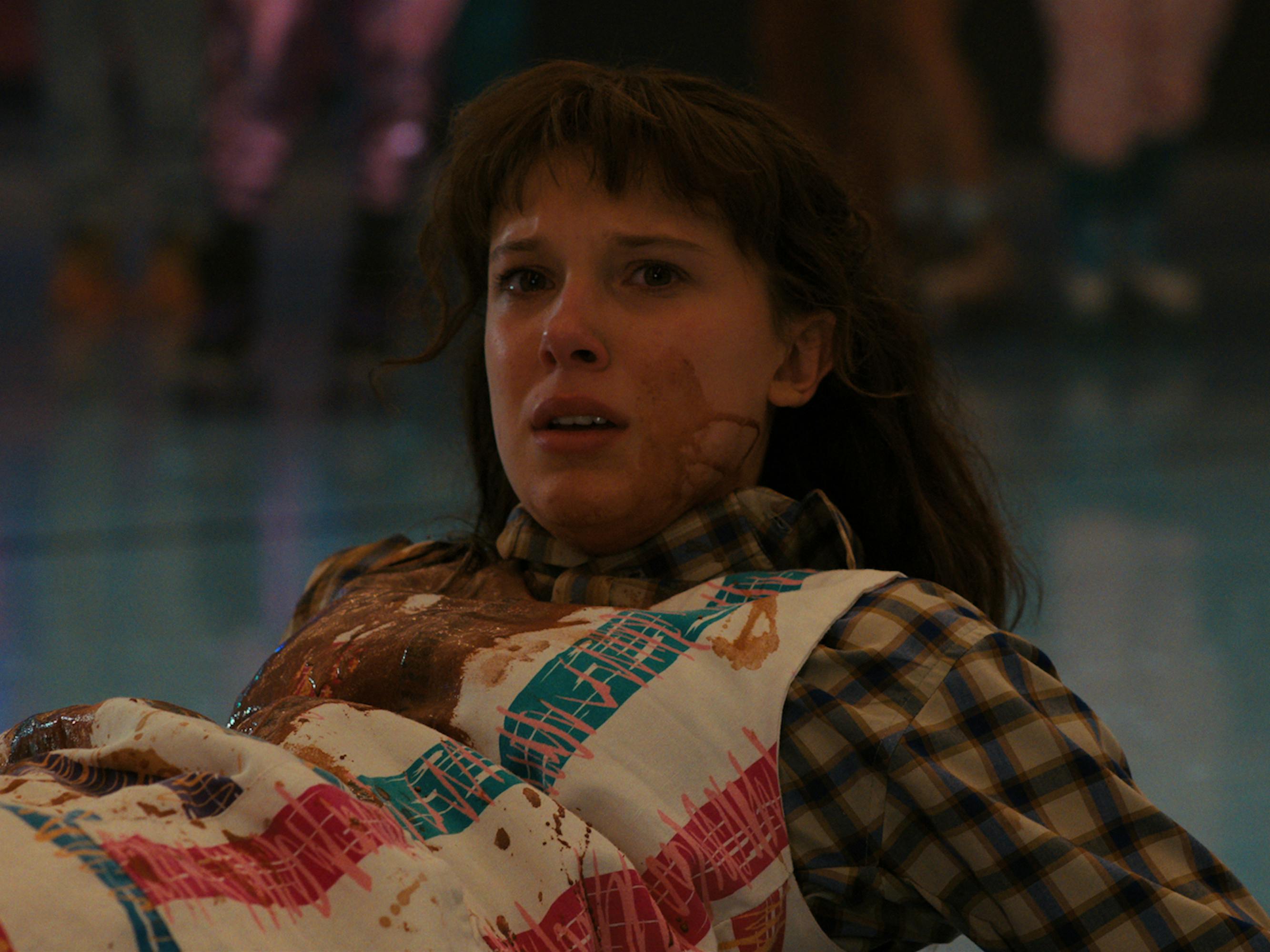 Eleven (Millie Bobby Brown) wears a checkered shirt under a food splattered dress and lies on the ground.
