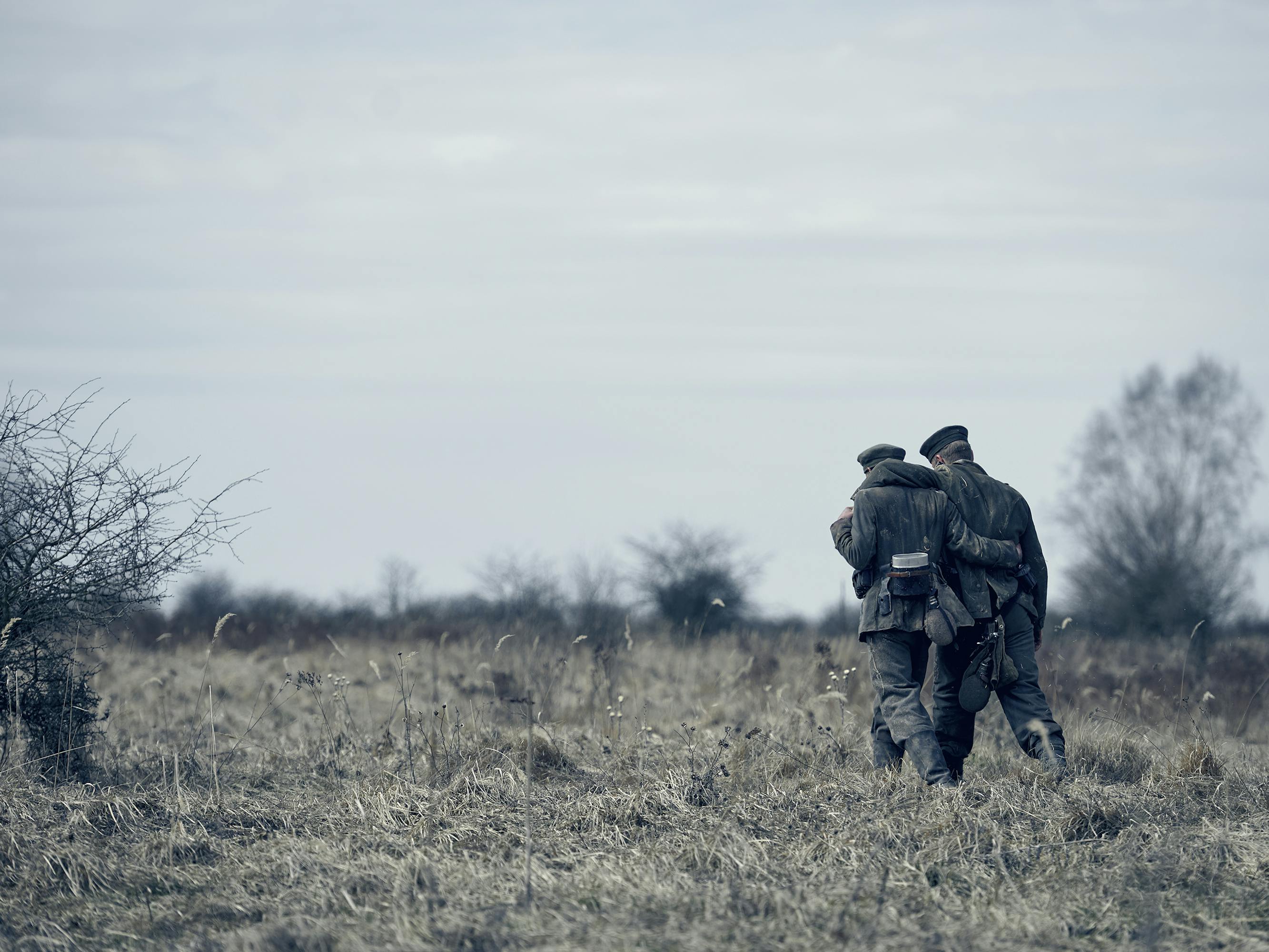 Two cast members walk through a grey field with their arms around each others backs.