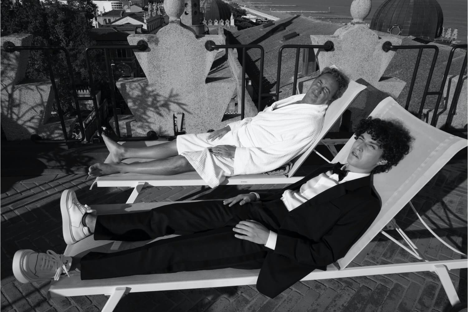 Paolo Sorretino and Filippo Scotti lounge on chaise lounges on a small porch. Sorrentino wears a robe, and Scotti wears a suit with sneakers.