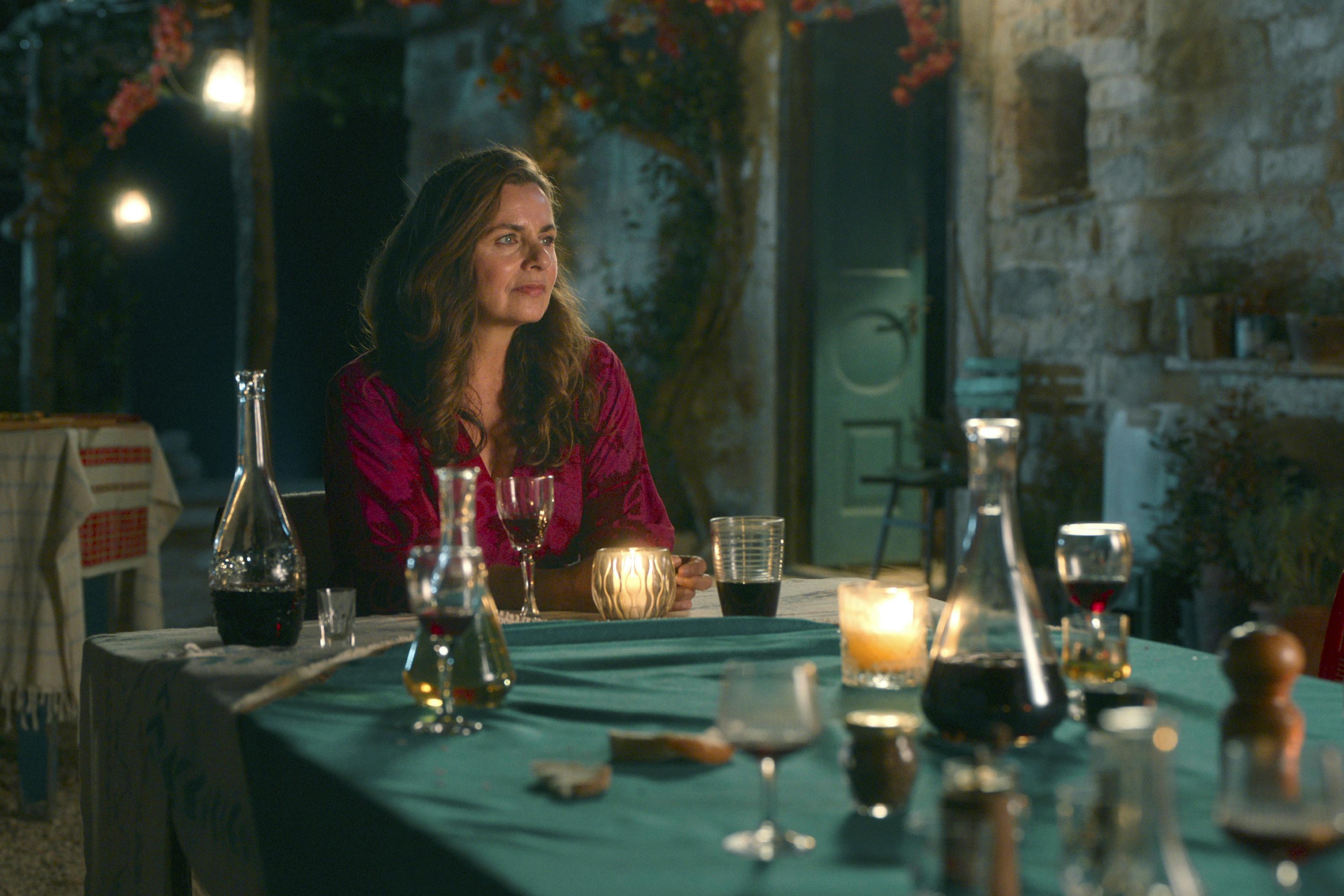 Zeynep (Naomi Krauss) sits at a table with wine and olive oil in front of her. 