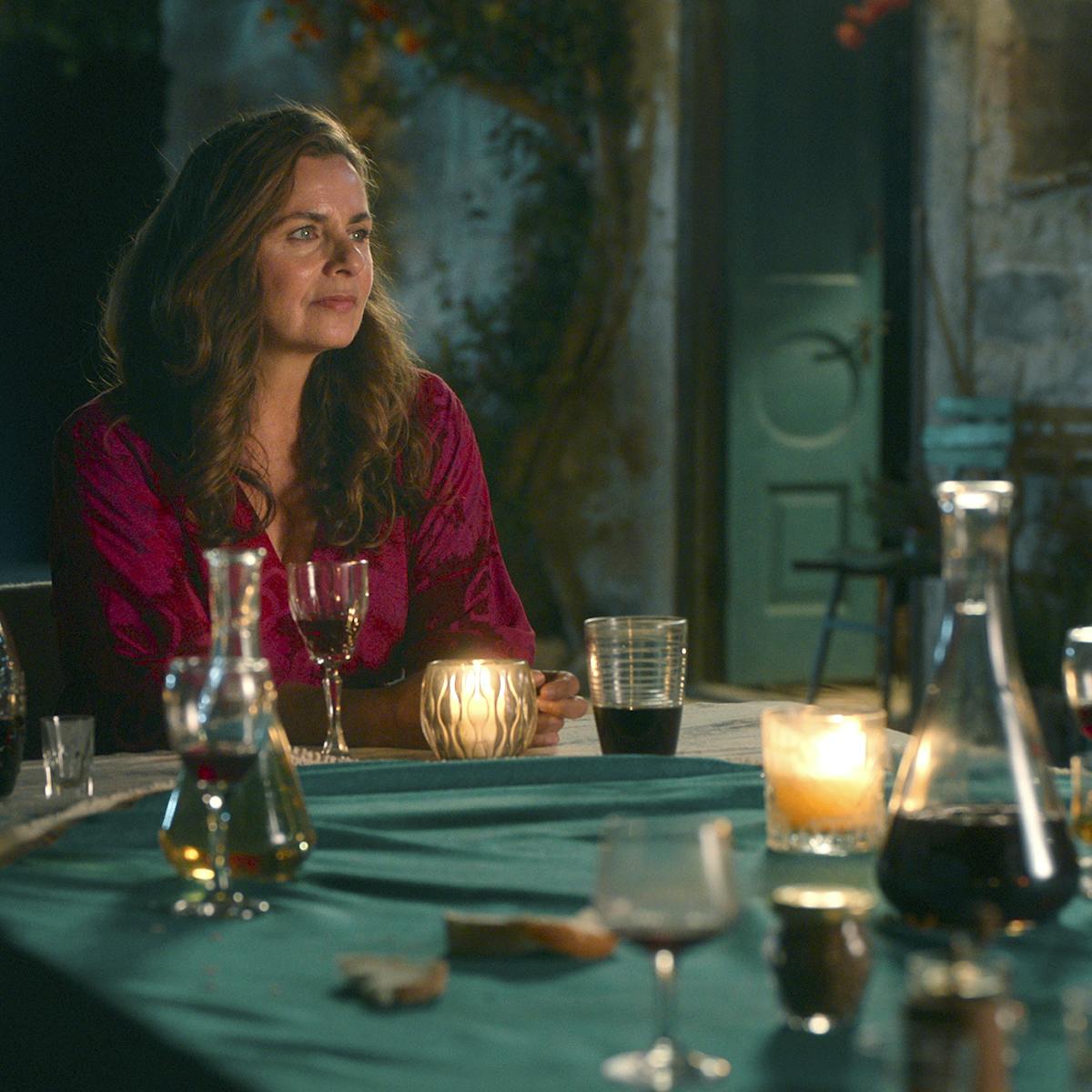 Zeynep (Naomi Krauss) sits at a table with wine and olive oil in front of her. 