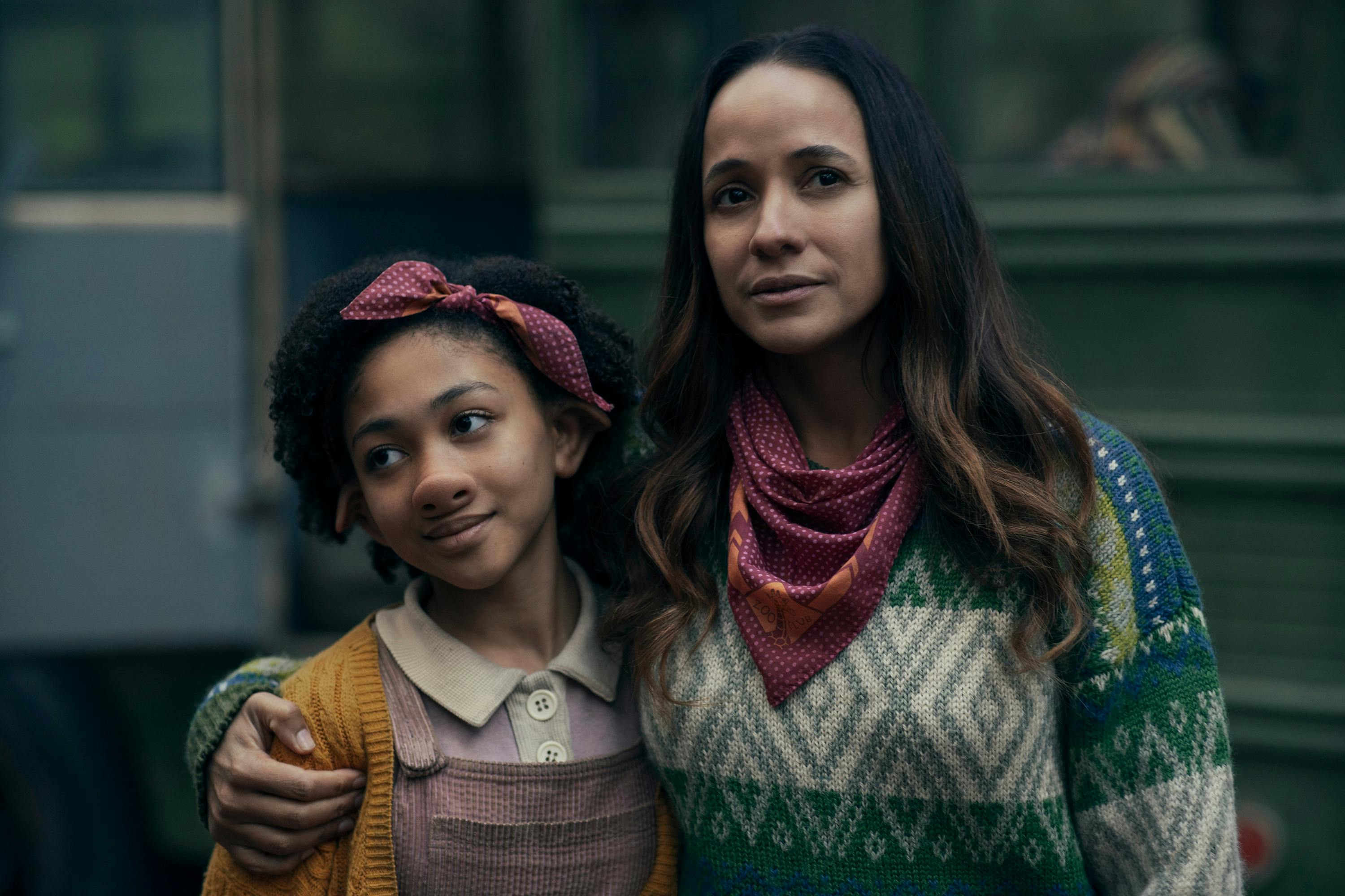 Wendy (Naledi Murray) and Aimee (Dania Ramirez) stand together outside. Wendy wears purple corduroy overalls and a yellow cardigan, and a headband. Aimee wears a patterned sweater and a scarf with the same pattern as Wendy’s headband around her neck. 
