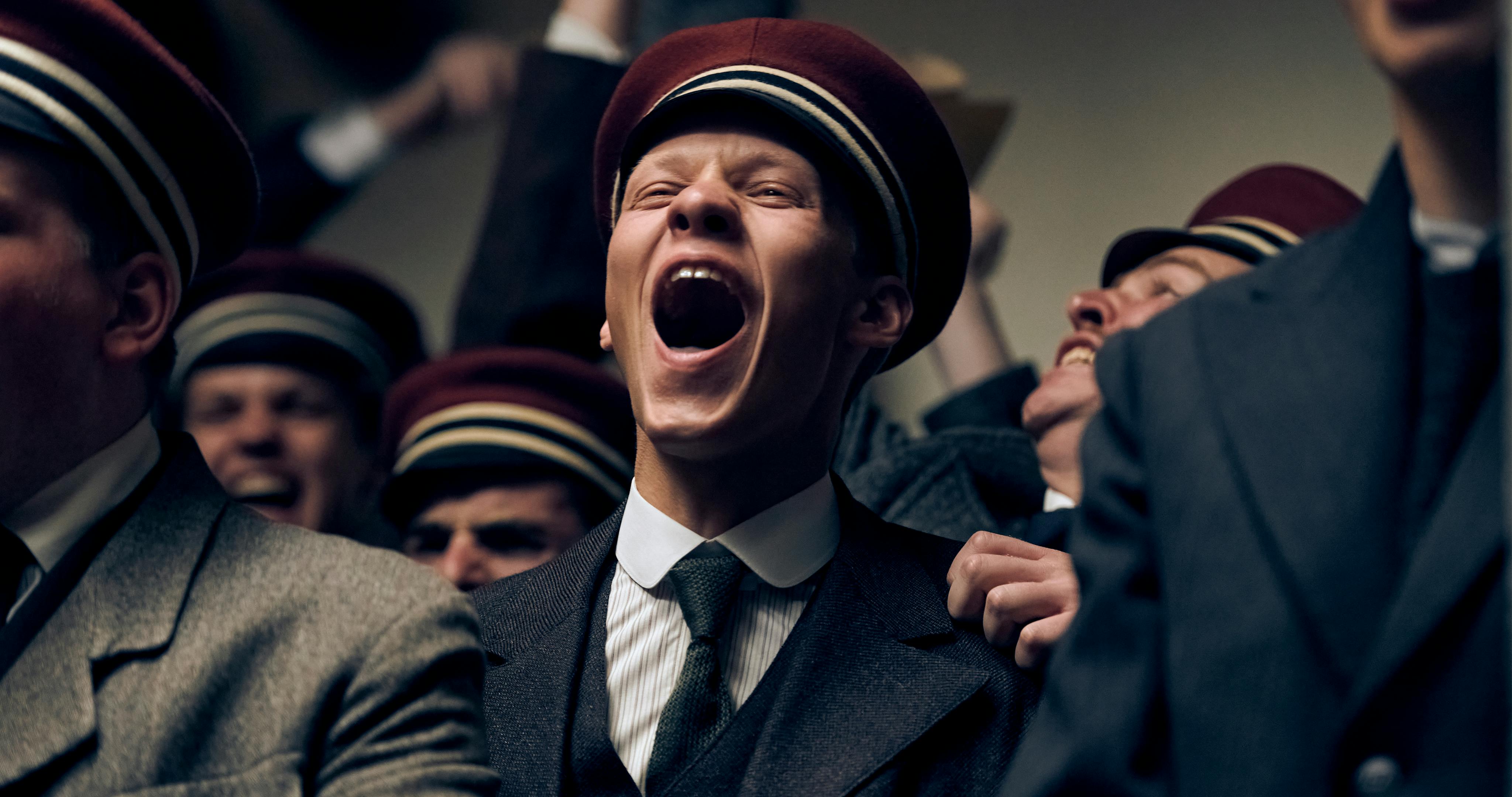 Paul Bäumer (Felix Kammerer) wears a black suit and tie and a red hat. His mouth is wide open as he cheers, in a pack of other cheering men. 