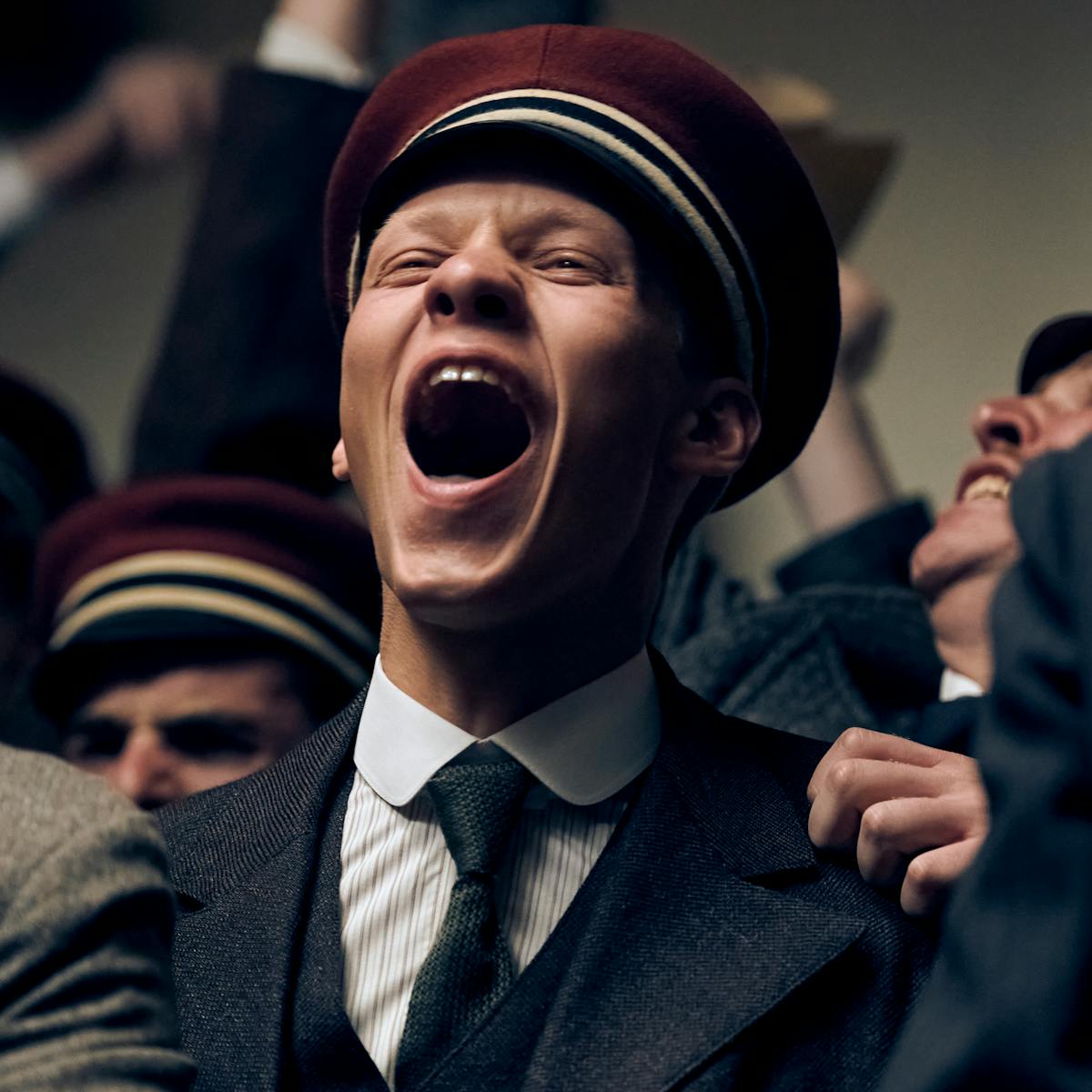 Paul Bäumer (Felix Kammerer) wears a black suit and tie, and a red hat. His mouth is wide open as he cheers, in a pack of other cheering men. 