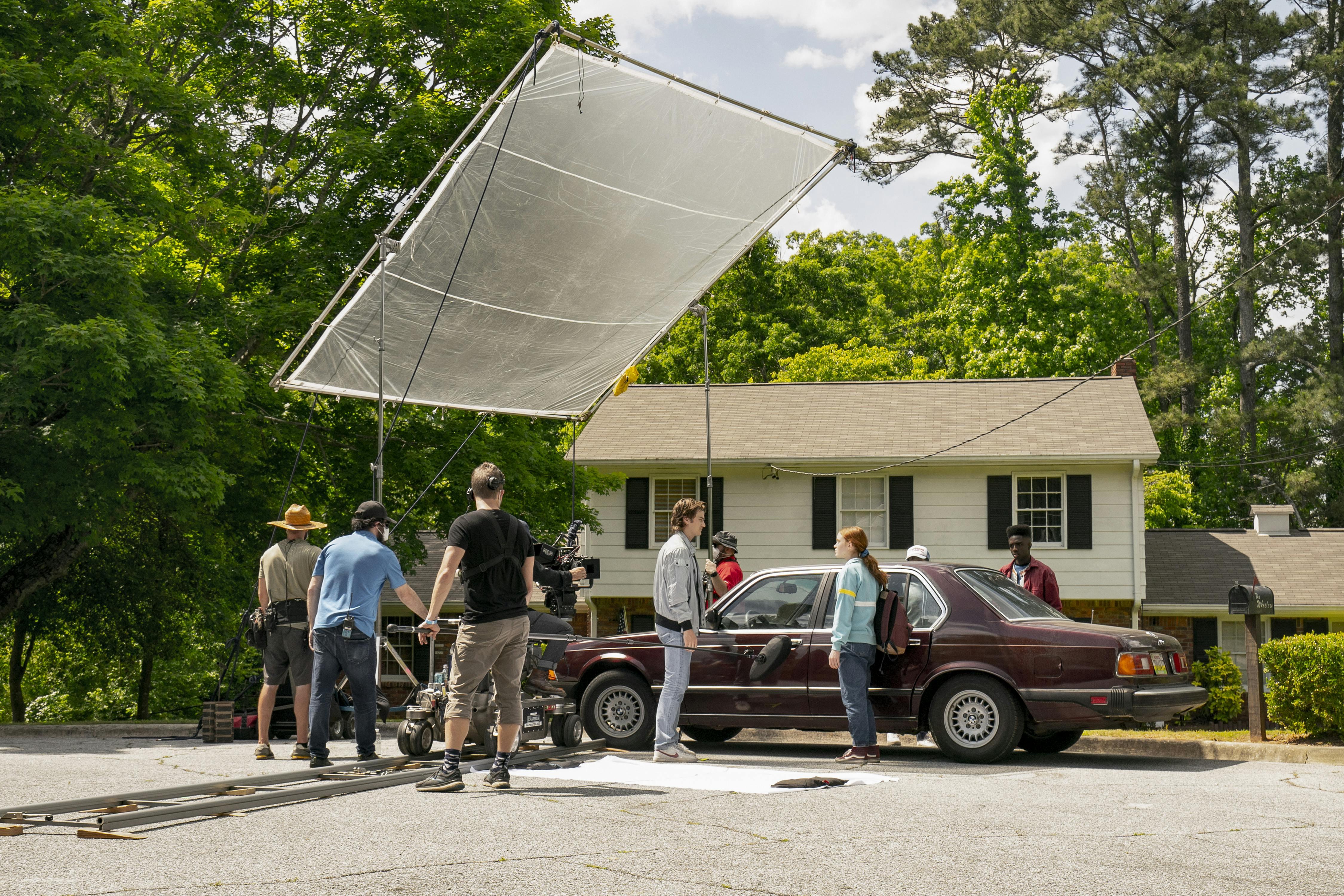 Cast and crew work outside. There is a huge reflective screen, a maroon car, and a white house. The white picket fence scene is complete with greenery and a blue sky.
