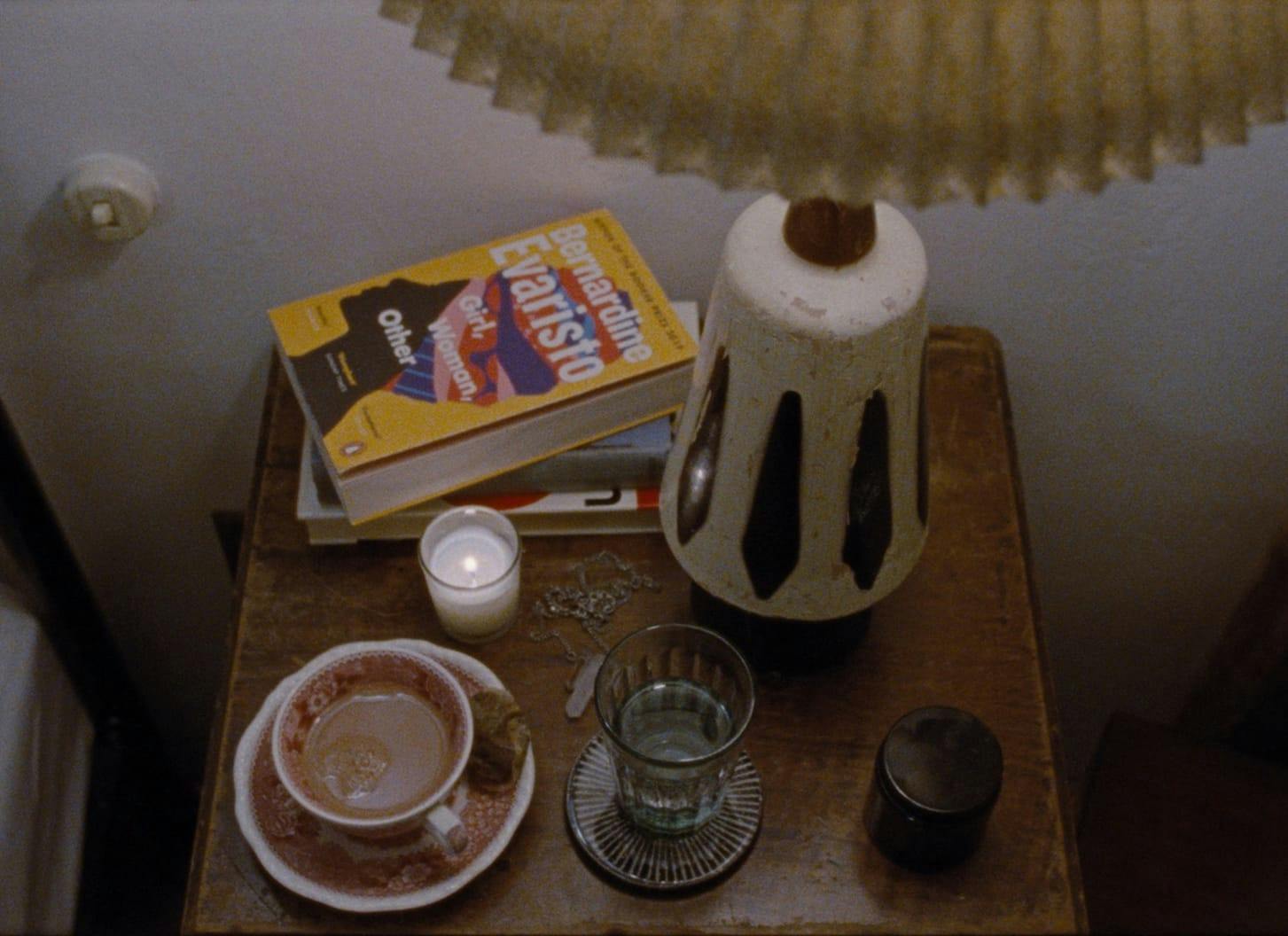 A shot of a table top complete with mug of coffee, glass of water, grinder, books, candle and lamp.