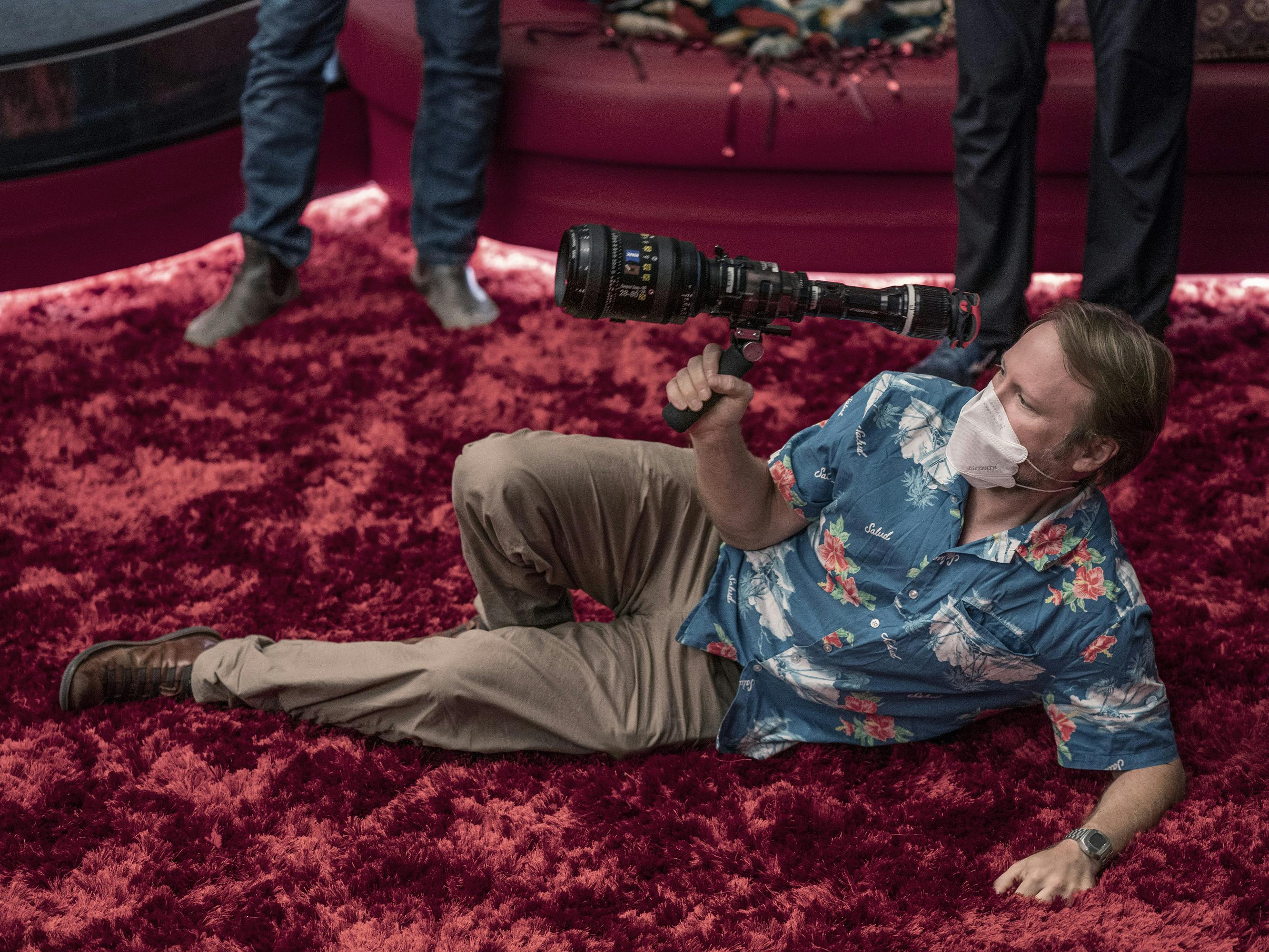 Rian Johnson wears a Hawaiian shirt and khaki pants and lies on a plush, red rug holding a lens. 