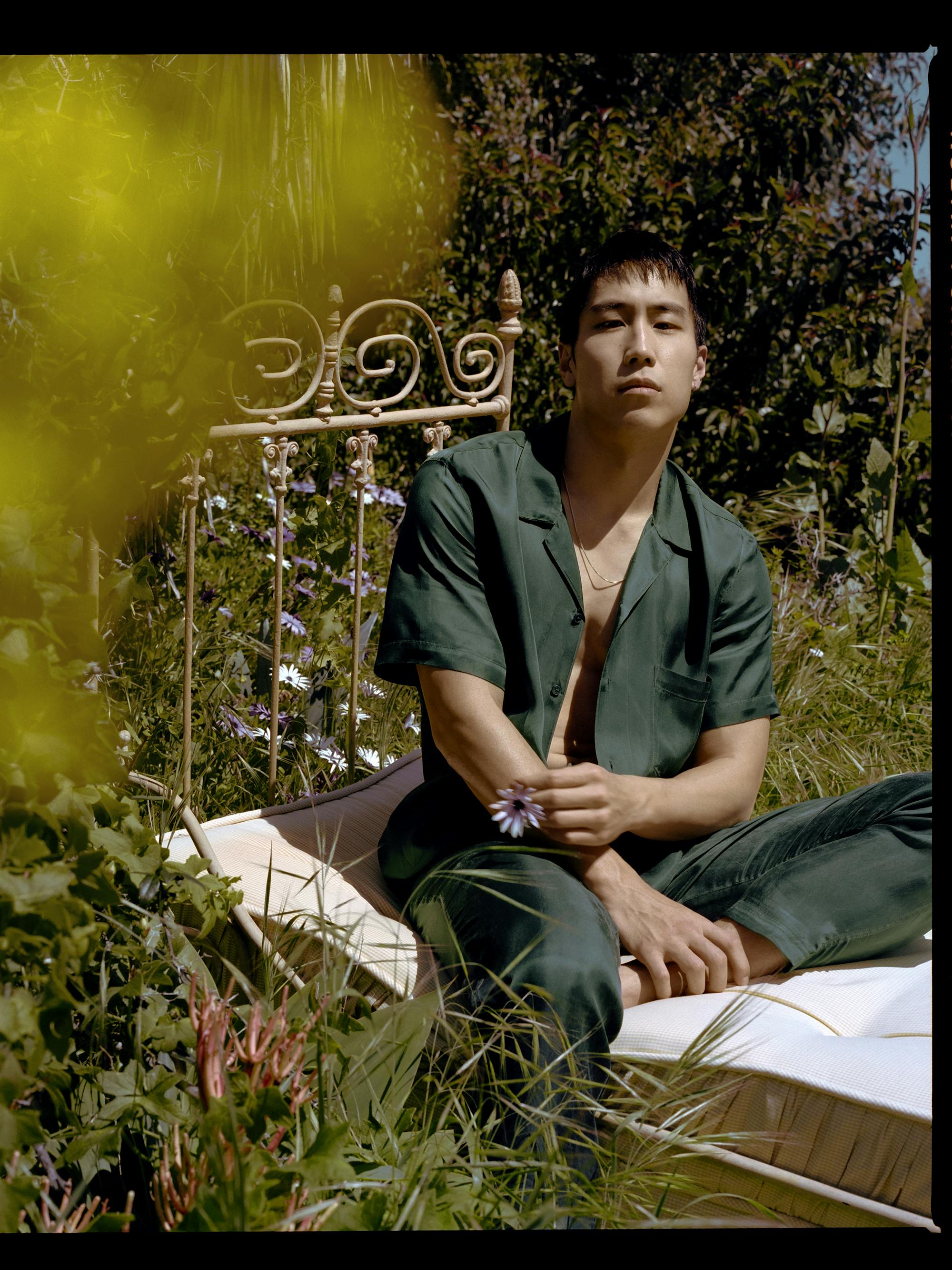 Young Mazino wears a green jumpsuit and sits on a metal bed in a field of grass and flowers.