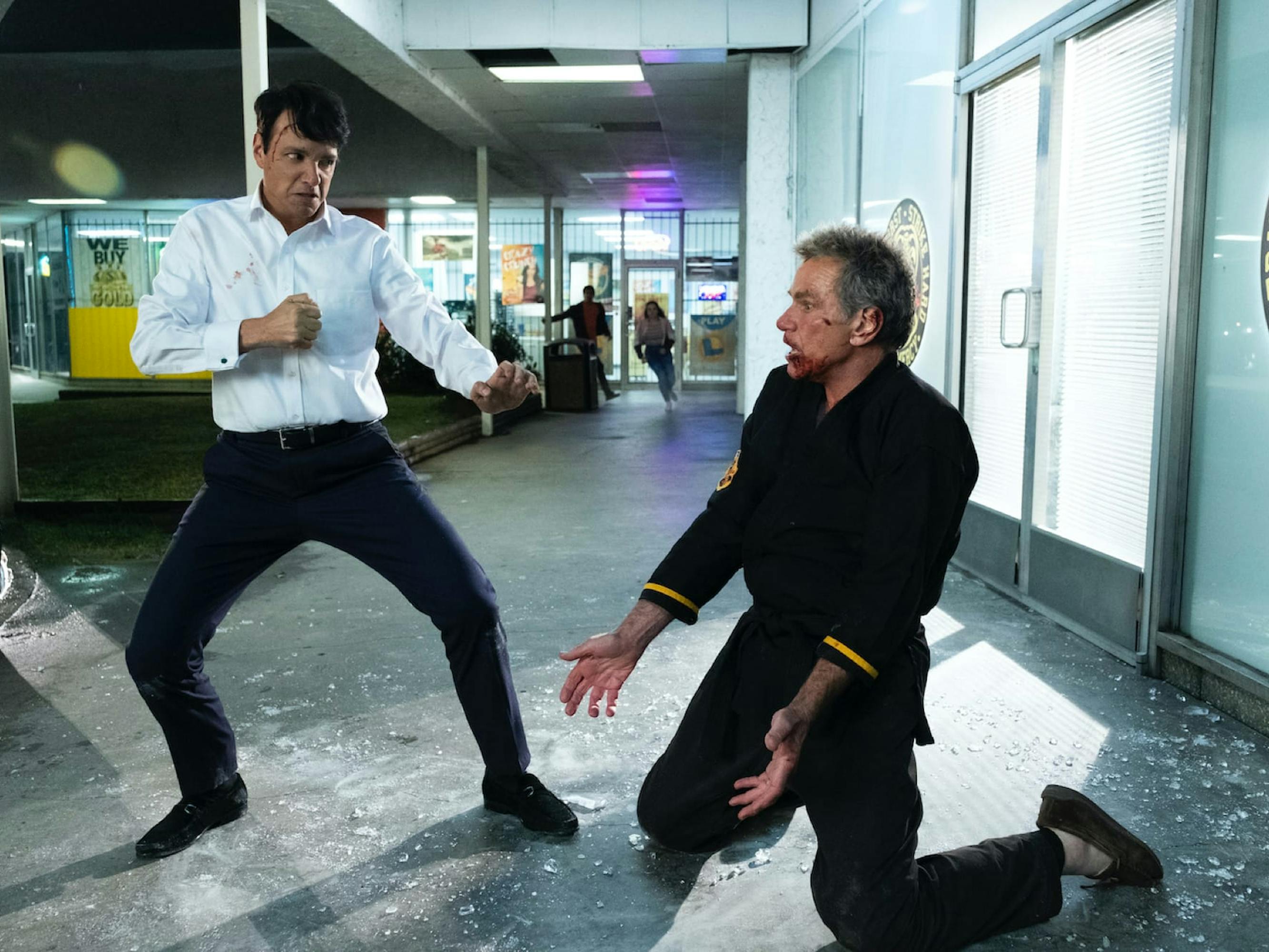 Daniel LaRusso (Ralph Macchio) and John Kreese (Martin Kove) stand off in a fight in a car dealership. LaRusso wears a white shirt and stands ready to strike. Kreese kneels on the ground in a pile of glass, bloodied.