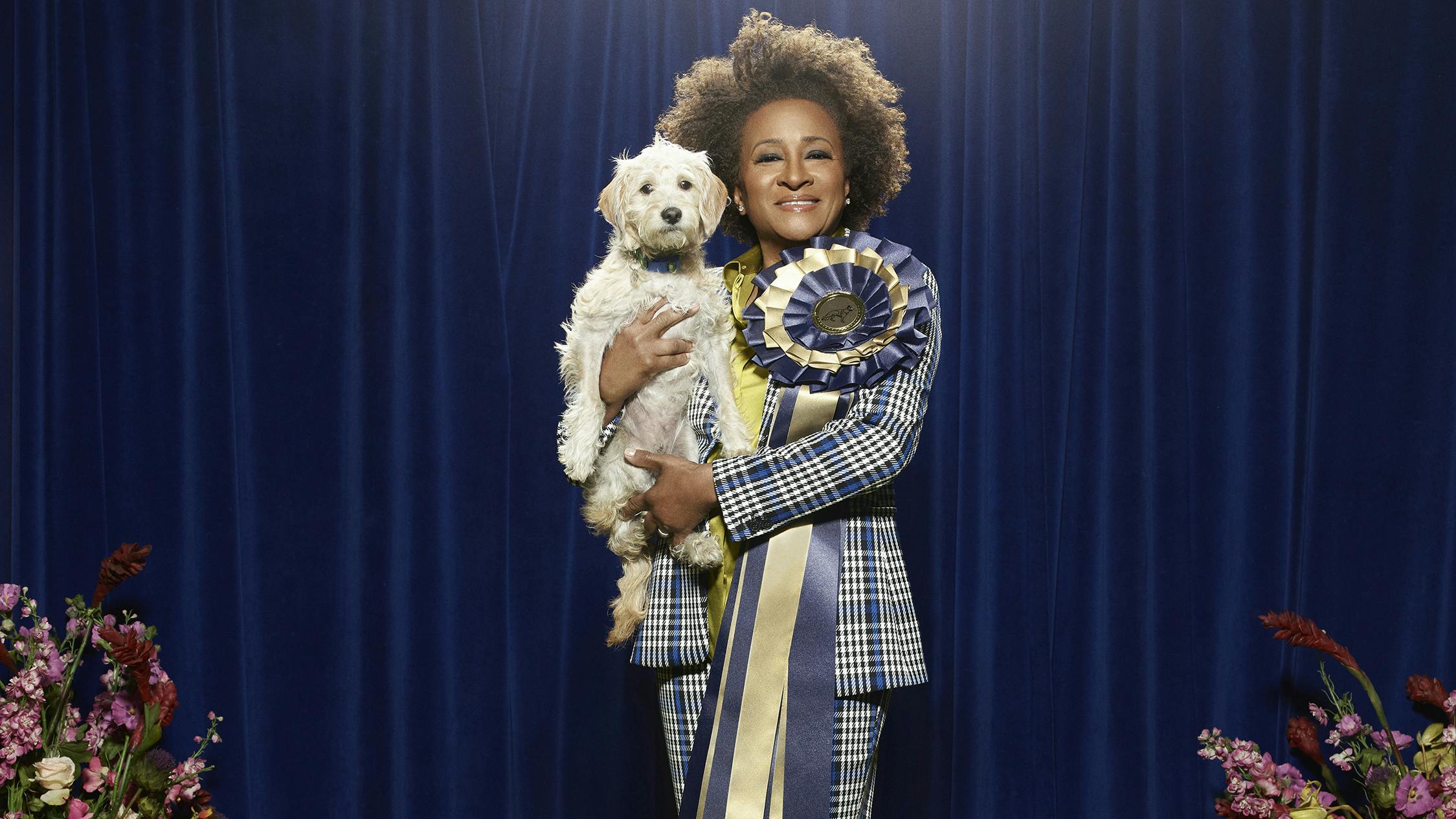 Wanda Sykes wears a plaid suit and holds a small dog. Pinned to her lapel is a gold and blue ribbon.