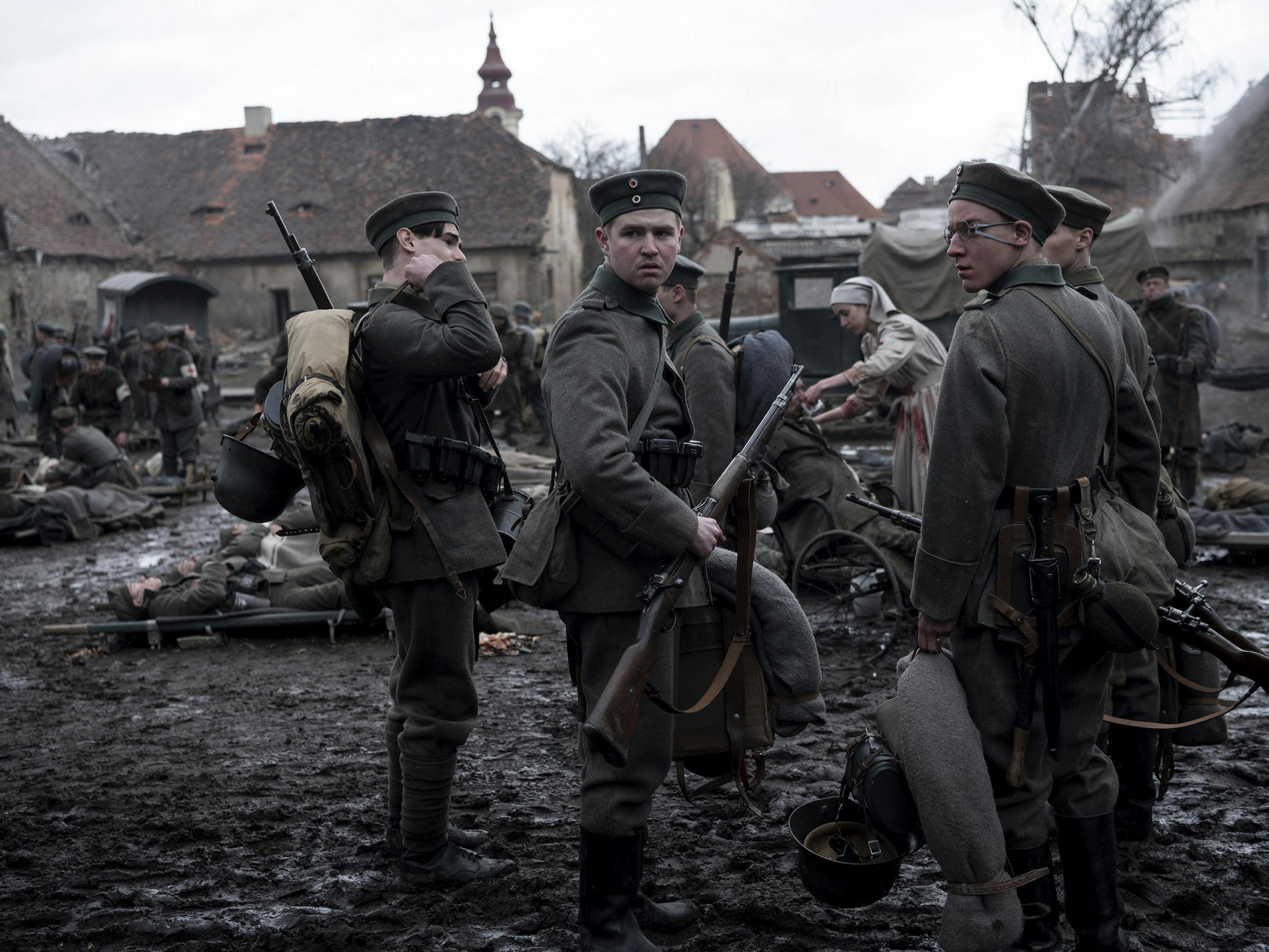 Albert Kropp (Aaron Hilmer), Franz Müller (Moritz Klaus), Lugwig Behm (Adrian Grünewald) stand with other soldiers in this muddy outdoor scene. Behind them is a nurse tending to an injury.