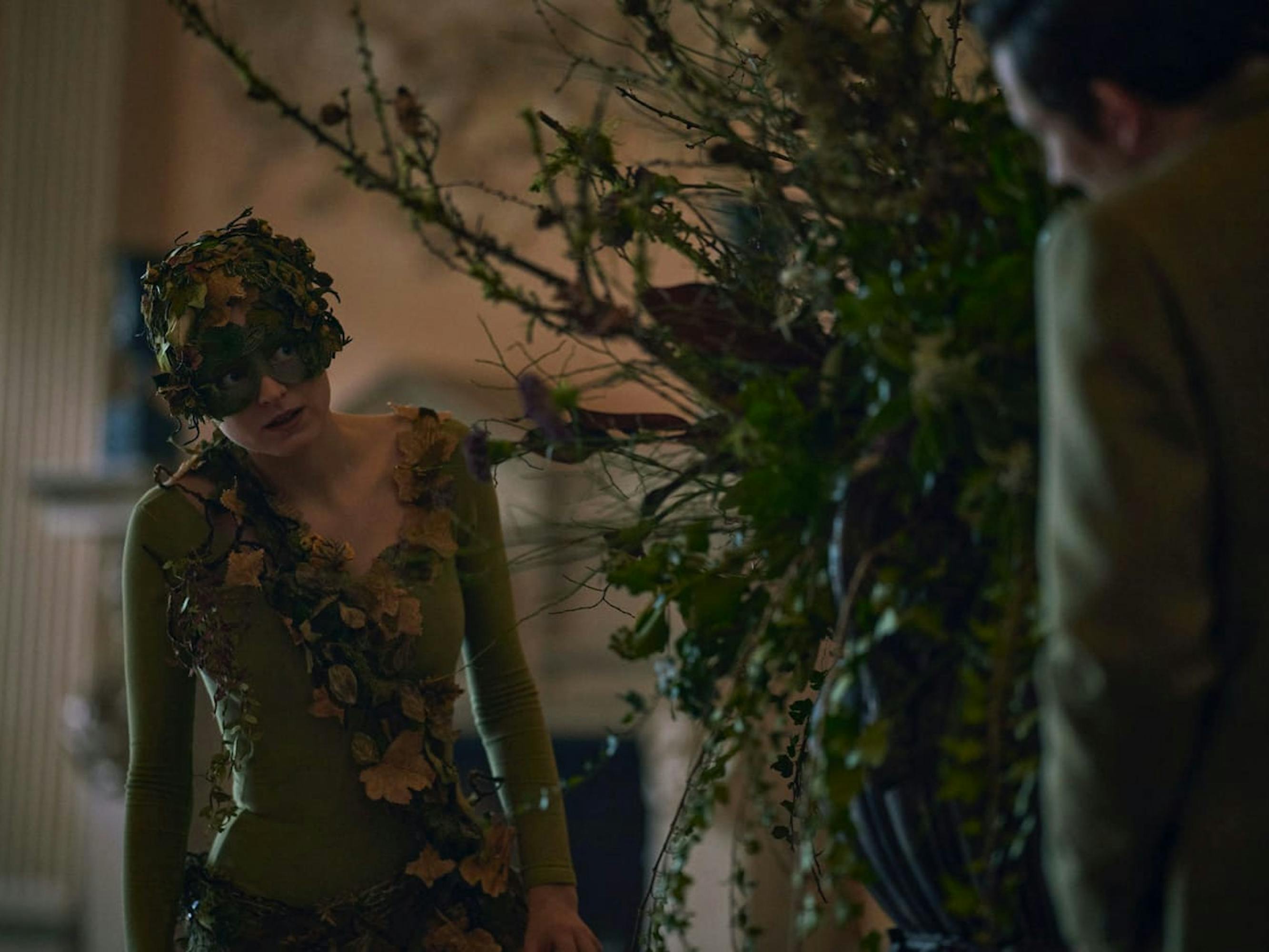 Diana (Emma Corrin) creepes behind a large planter, dressed as a tree from A Midsummer Night’s Dream. Josh’s figure appears on the right side.