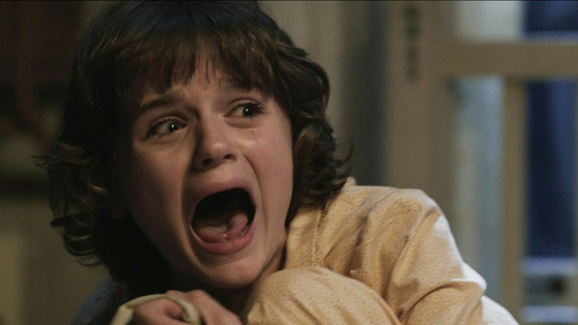 Christine (Joey King) in The Conjuring (2013). With her mouth open wide, she clutches her legs to her body in fear.