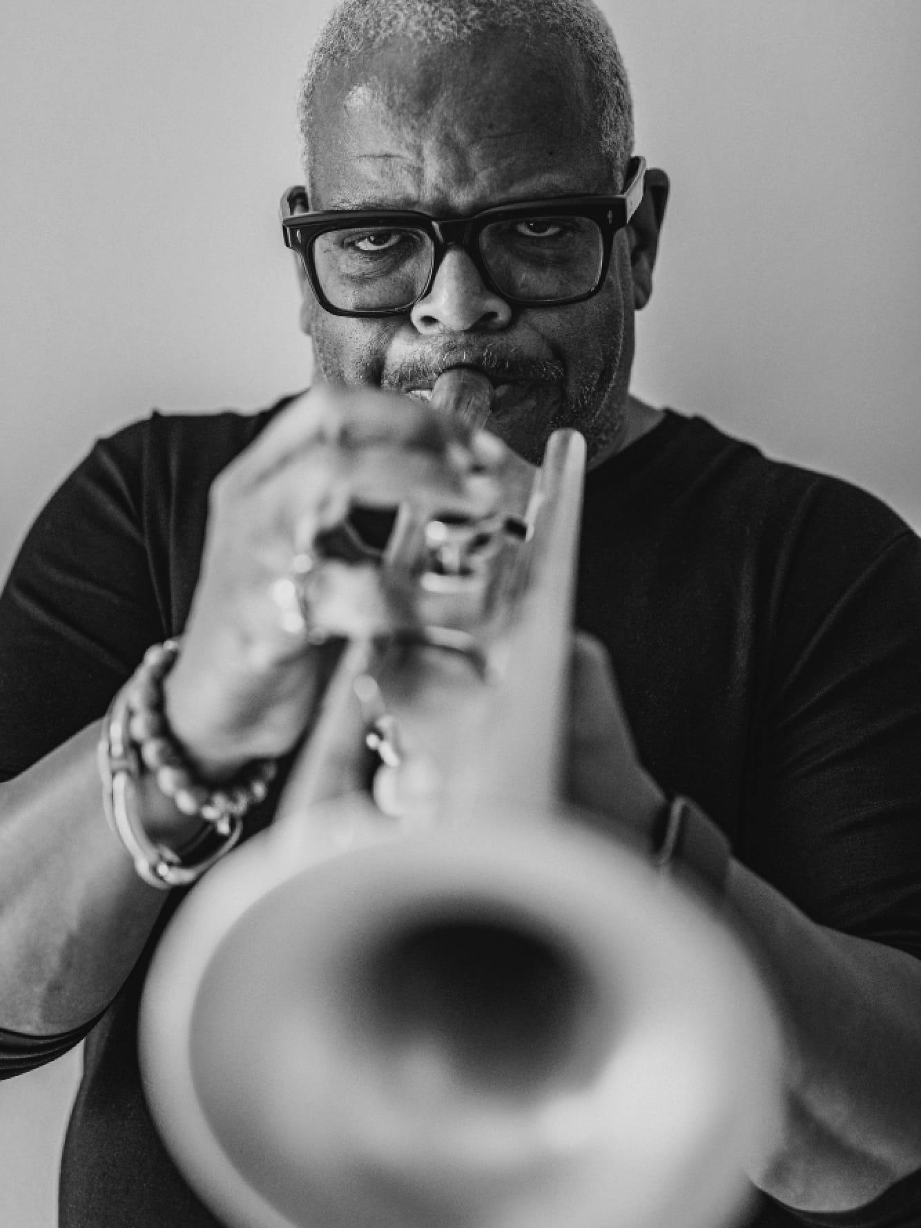 A black-and-white photograph of composer Terence Blanchard playing his trumpet. He wears square-rimmed glasses, a dark-colored shirt, and several rings and bracelets. He looks directly at the camera, and the bell of the trumpet stretches toward us, the focus blurring at the nearest point. 