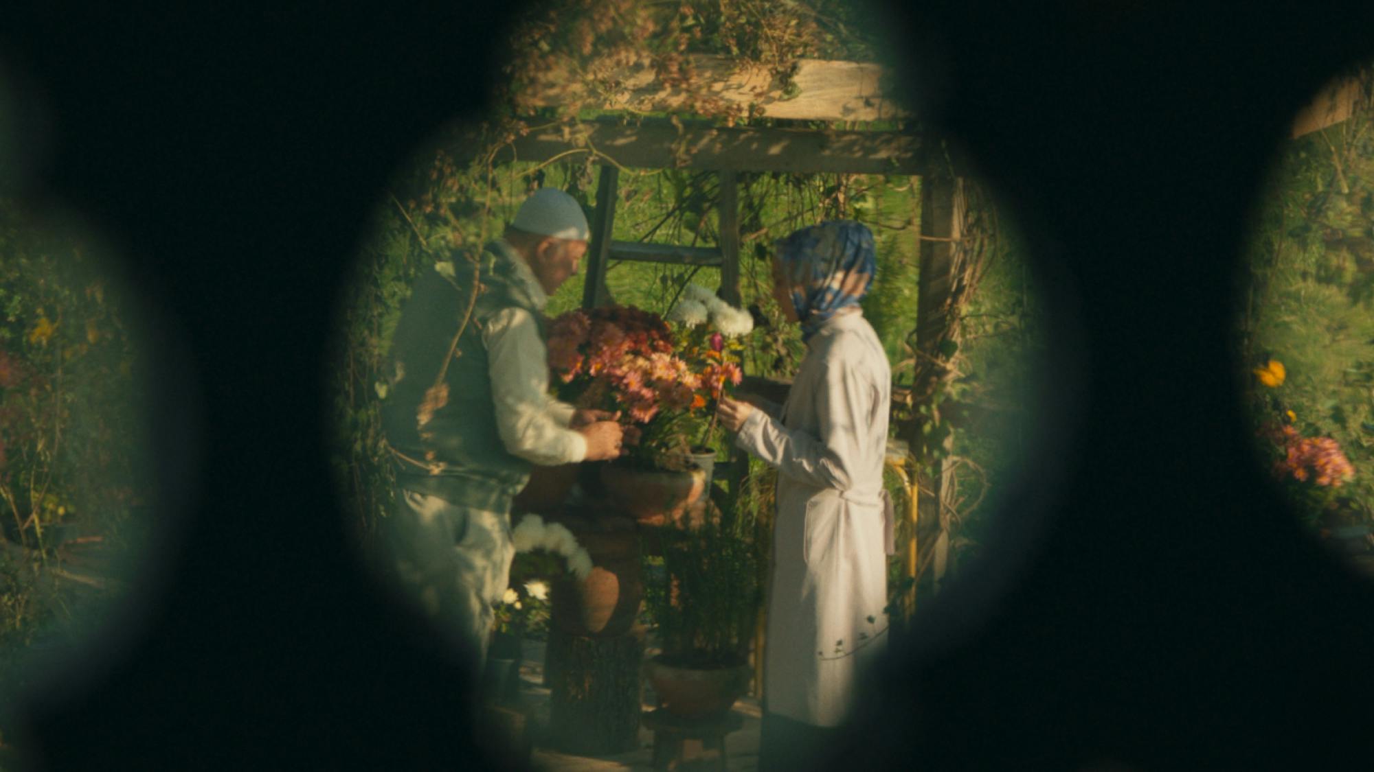 Meryem and her Hodja are seen through a fence, having a discussion over a planter of flowers in an idyllic green garden. 