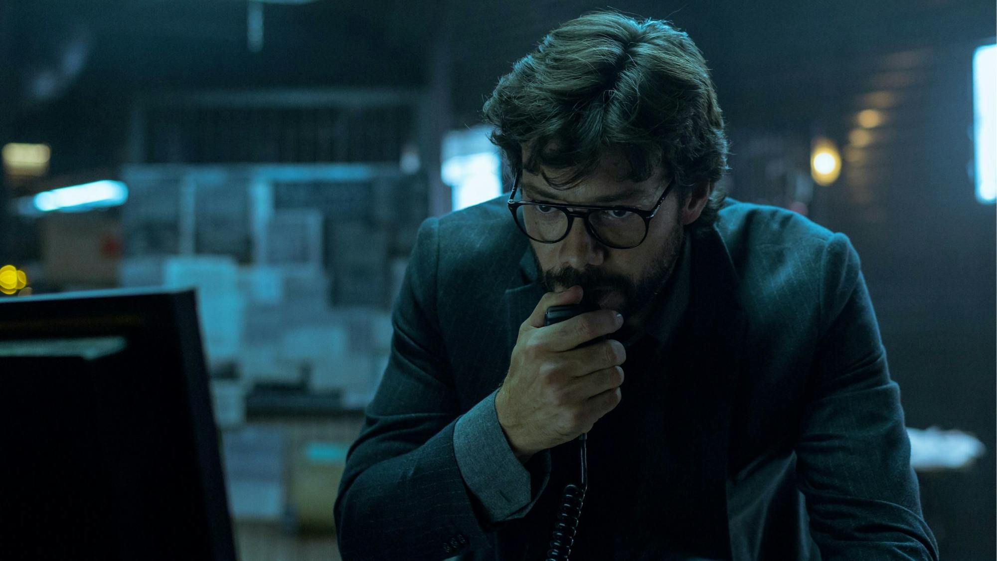 El Professor (Álvaro Morte) wears a dark suit and glasses and speaks into a radio. The mood in the room is ominous and grey.