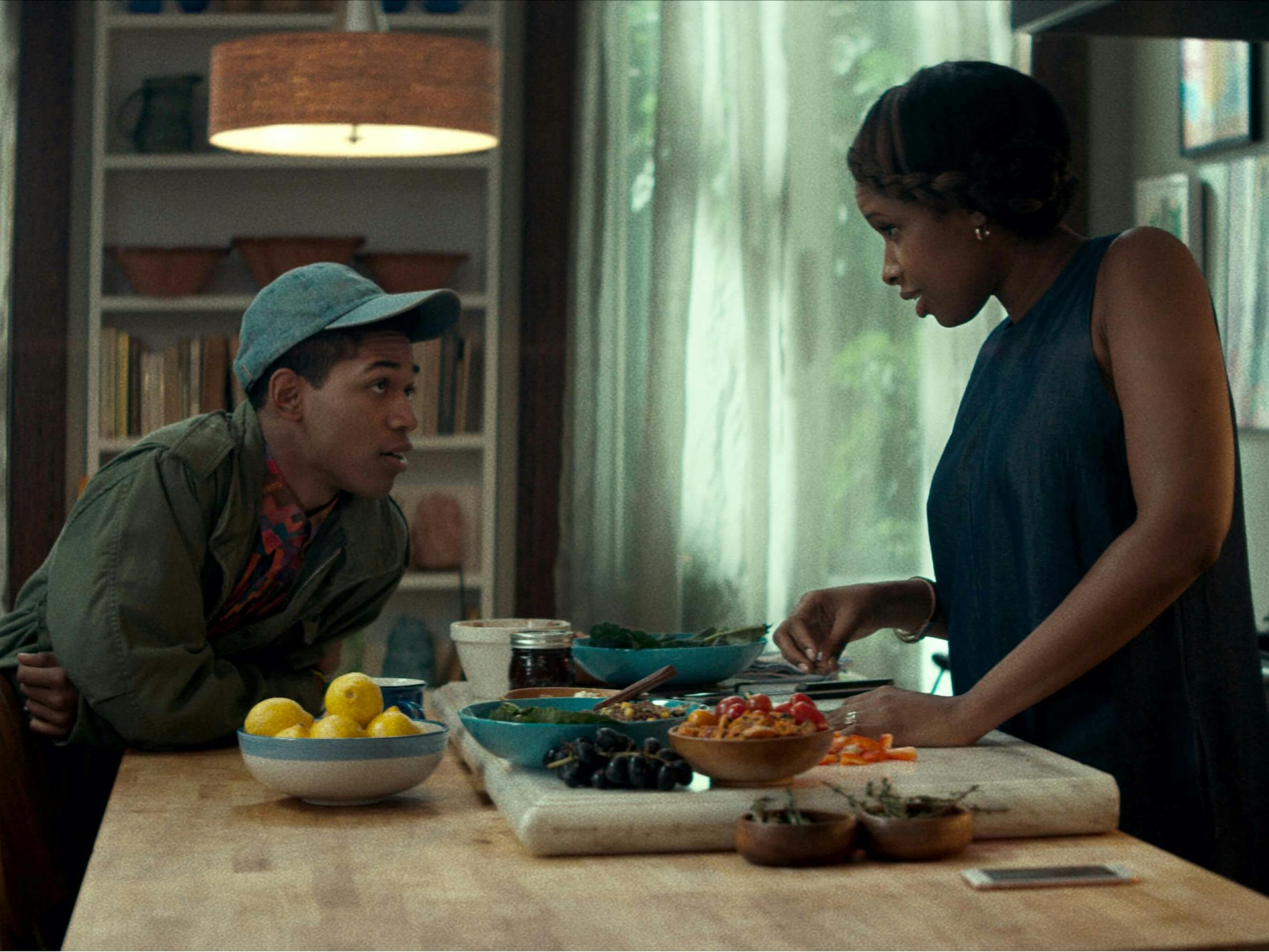 Harrison as Harmon in Monster. He wears a grey hat, green jacket, and red shirt. He leans across the kitchen table, adorned with fruit and lemons, to talk to his mom, played by Jennifer Hudson. She wears a navy sleeveless top.