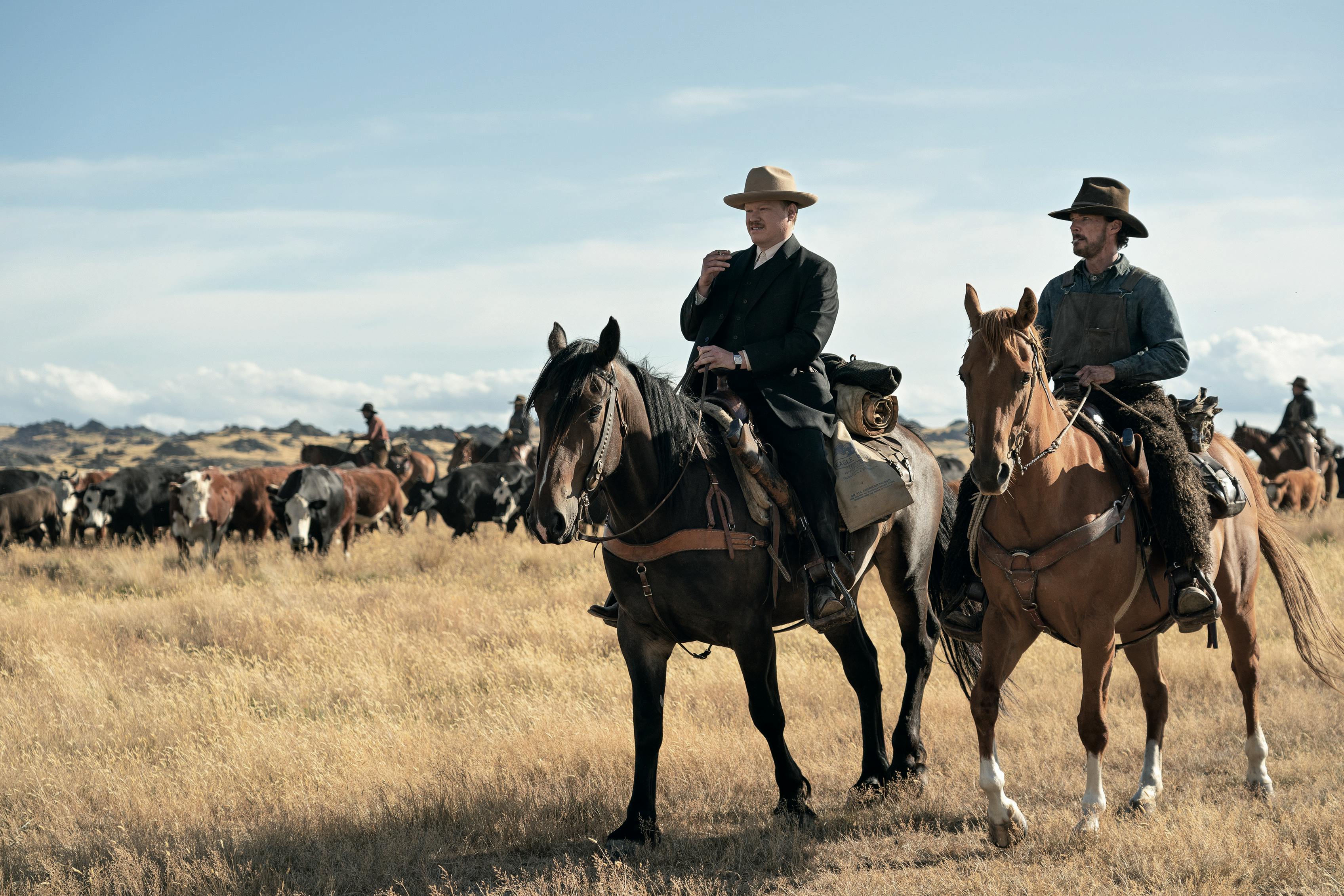 Jesse Plemons and Benedict Cumberbatch ride horses through a dry field. Behind them are tens of other horses.