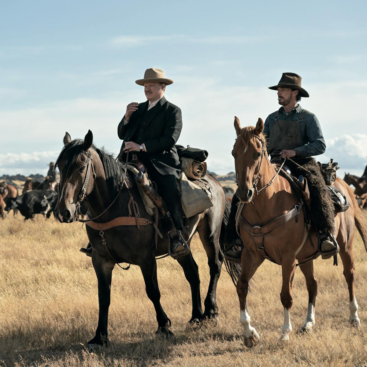 Jesse Plemons and Benedict Cumberbatch ride horses through a dry field. Behind them are tens of other horses.