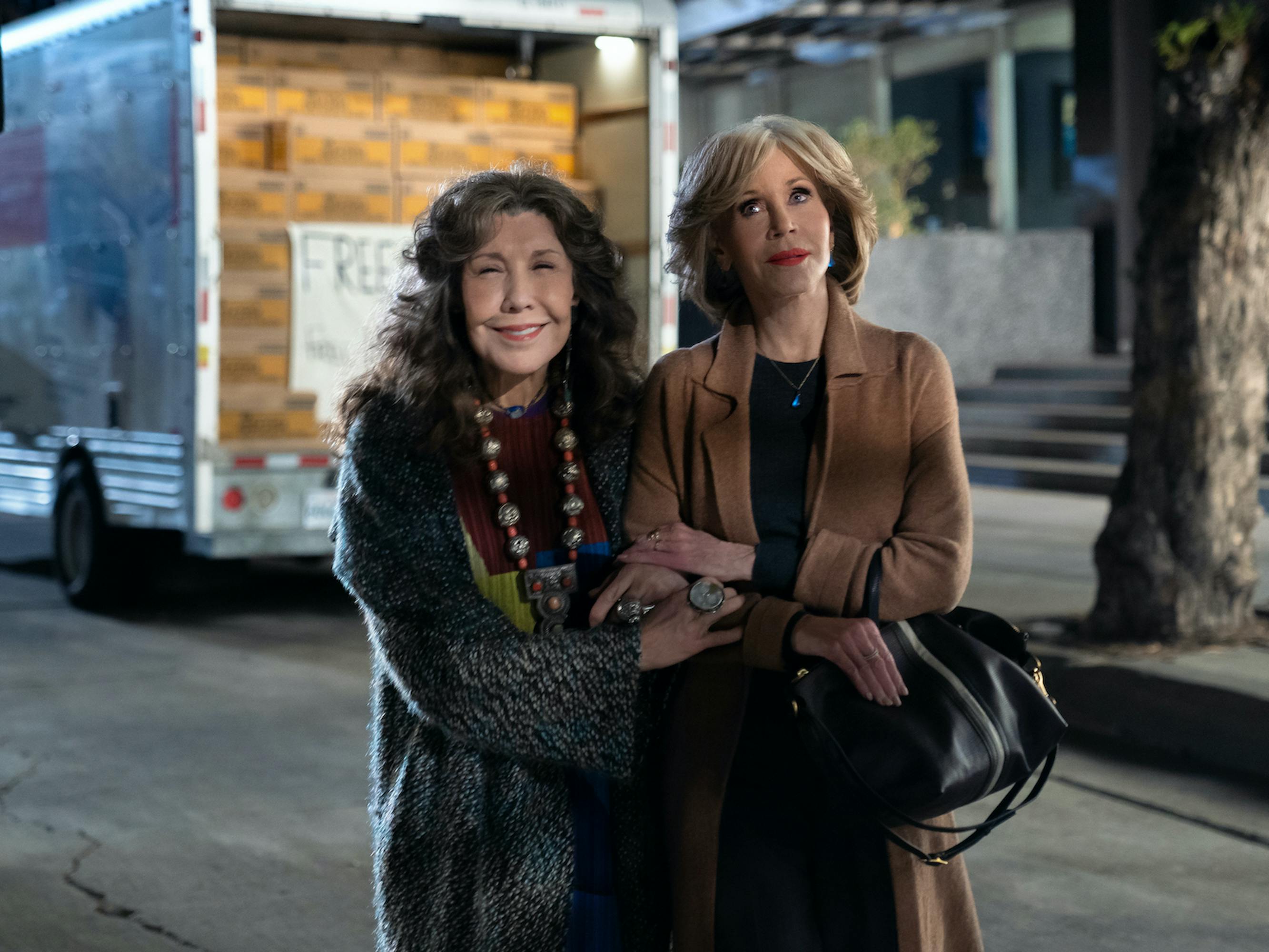 Frankie and Grace stand outside on a street. Frankie wears a shaggy grey jacket and a chunky necklace. Grace wears a red lip, camel jacket, black dress, and black bag. Behind them is a truck with boxes of something labeled 'Free.'