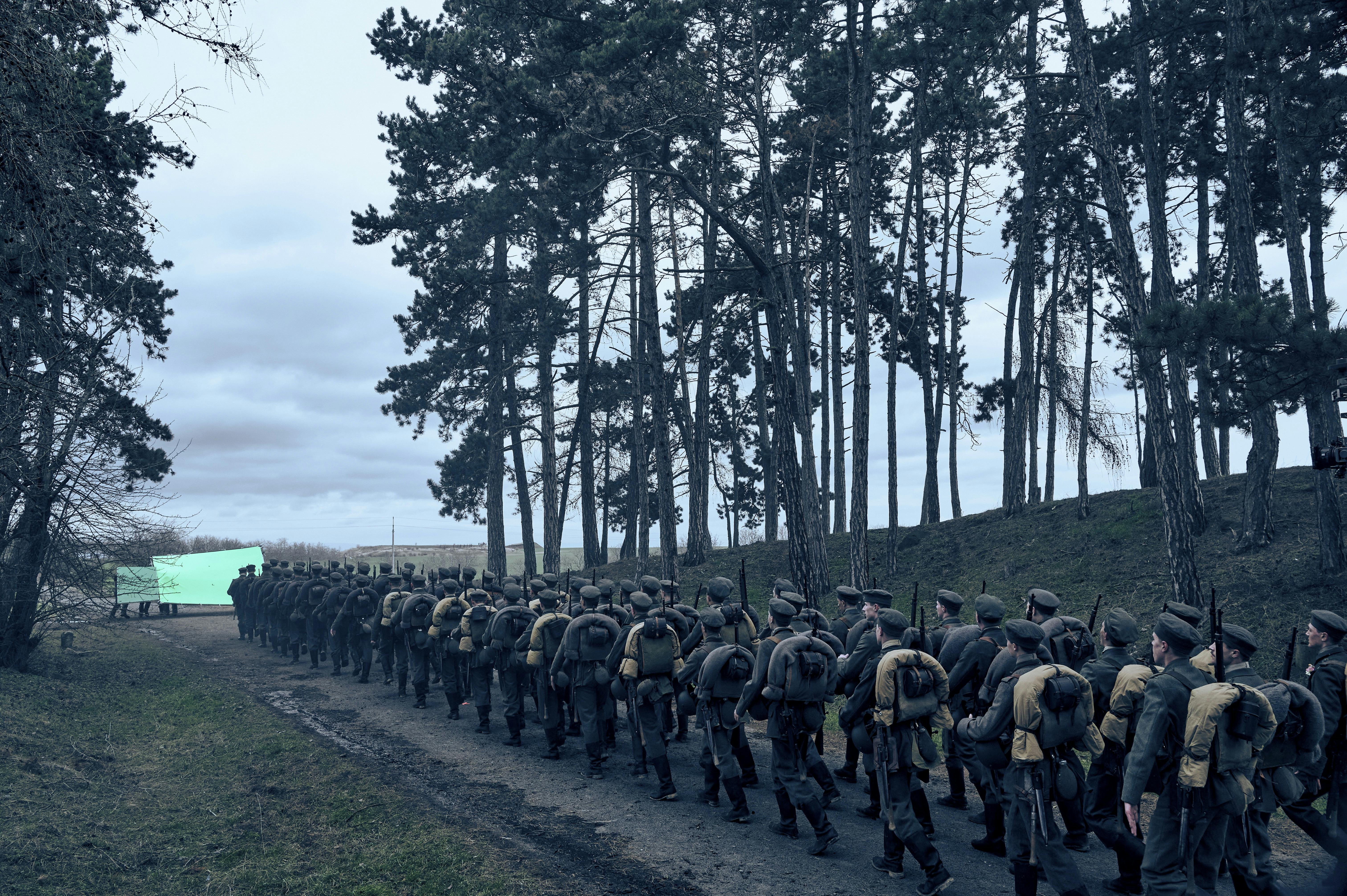 A line of soldiers stand together on a muddy path. Sparse trees line the path and the sky is green and cloudy.