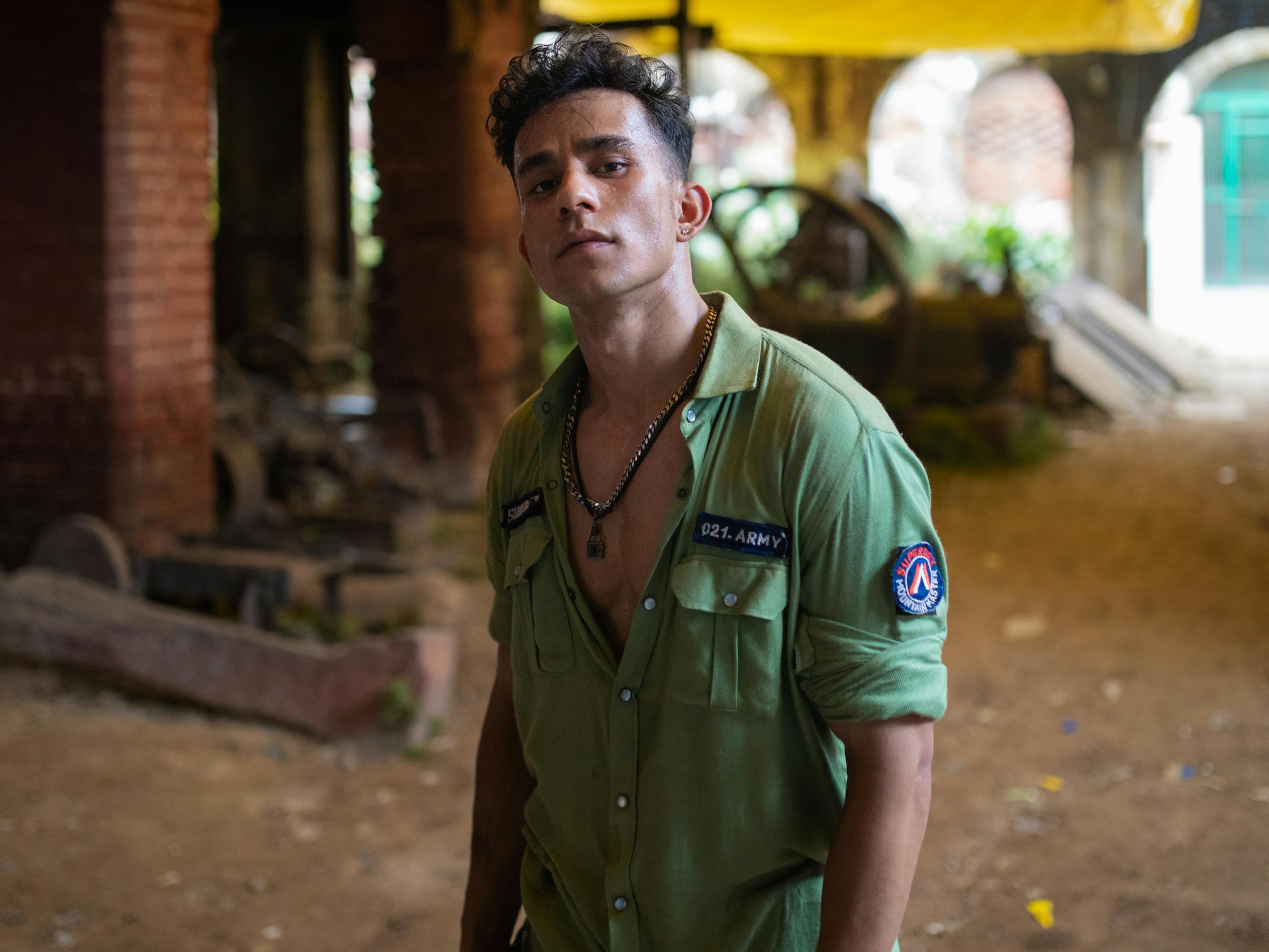 Balli (Cwaayal Sing) wears a mostly unbuttoned green shirt with patches and a dark necklace. 