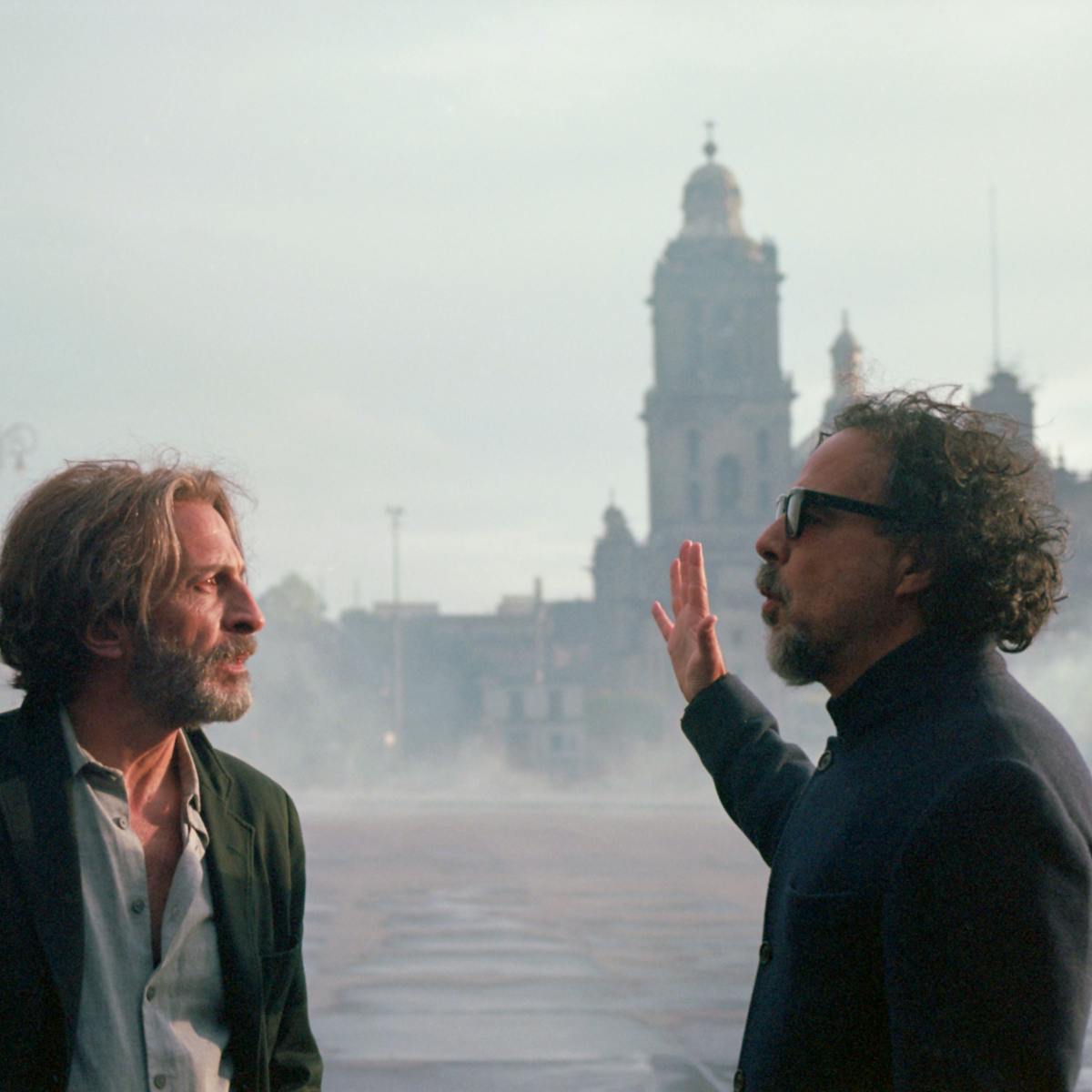 Daniel Giménez Cacho and Alejandro González Iñárritu talk outside an ornate building against a light grey sky.