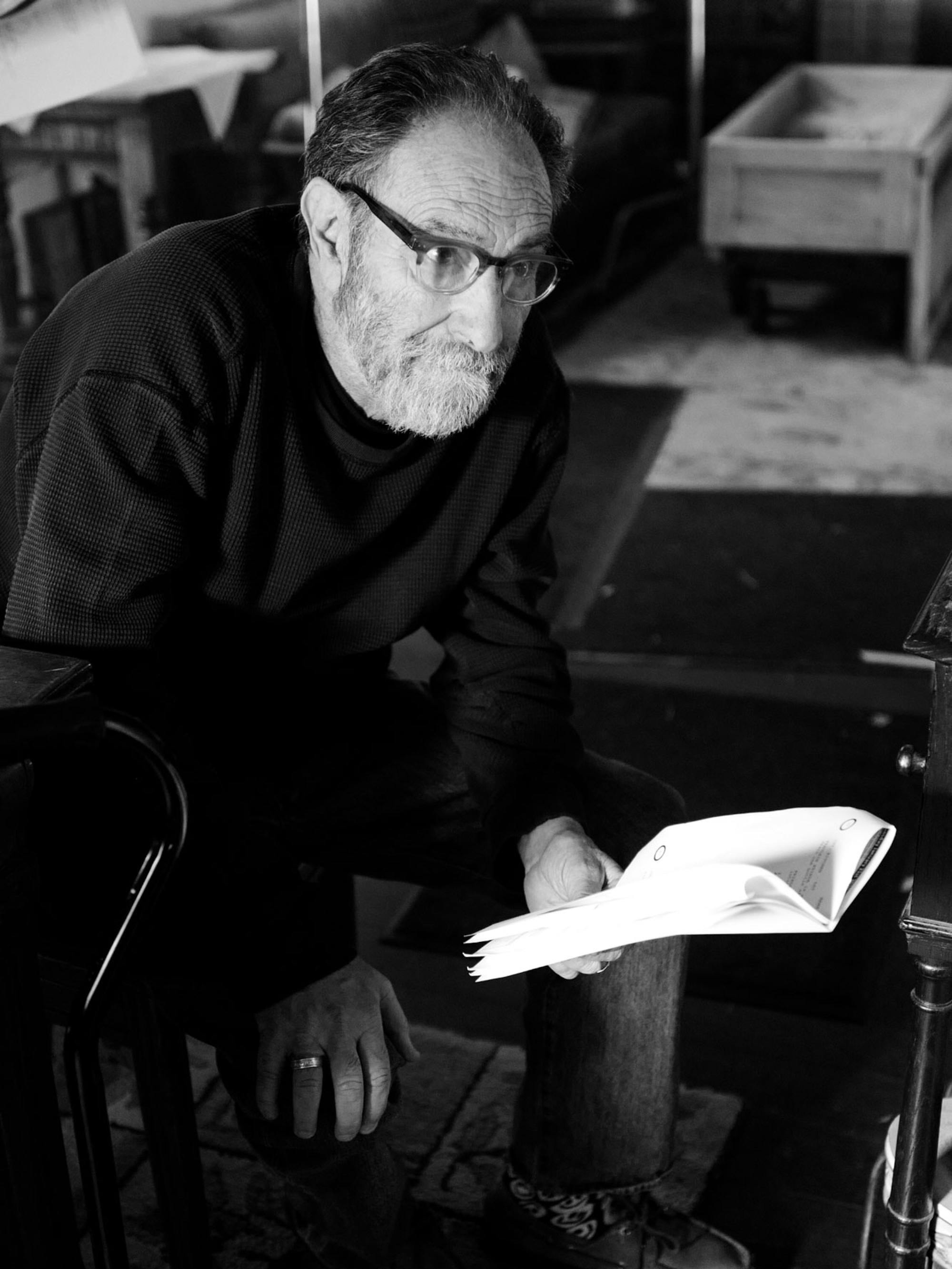 In this black-and-white photo, Roth, a white-bearded man with square-rimmed glasses, takes a knee and holds up a script. 