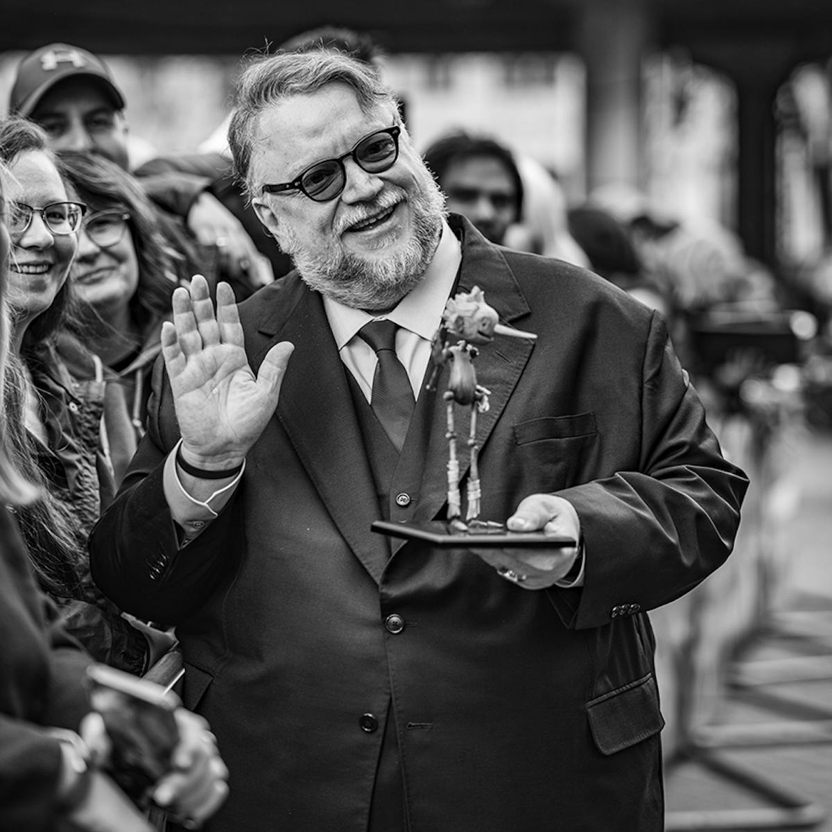 Guillermo del Toro wears a suit and waves from a crowd of people holding a miniature Pinocchio puppet.