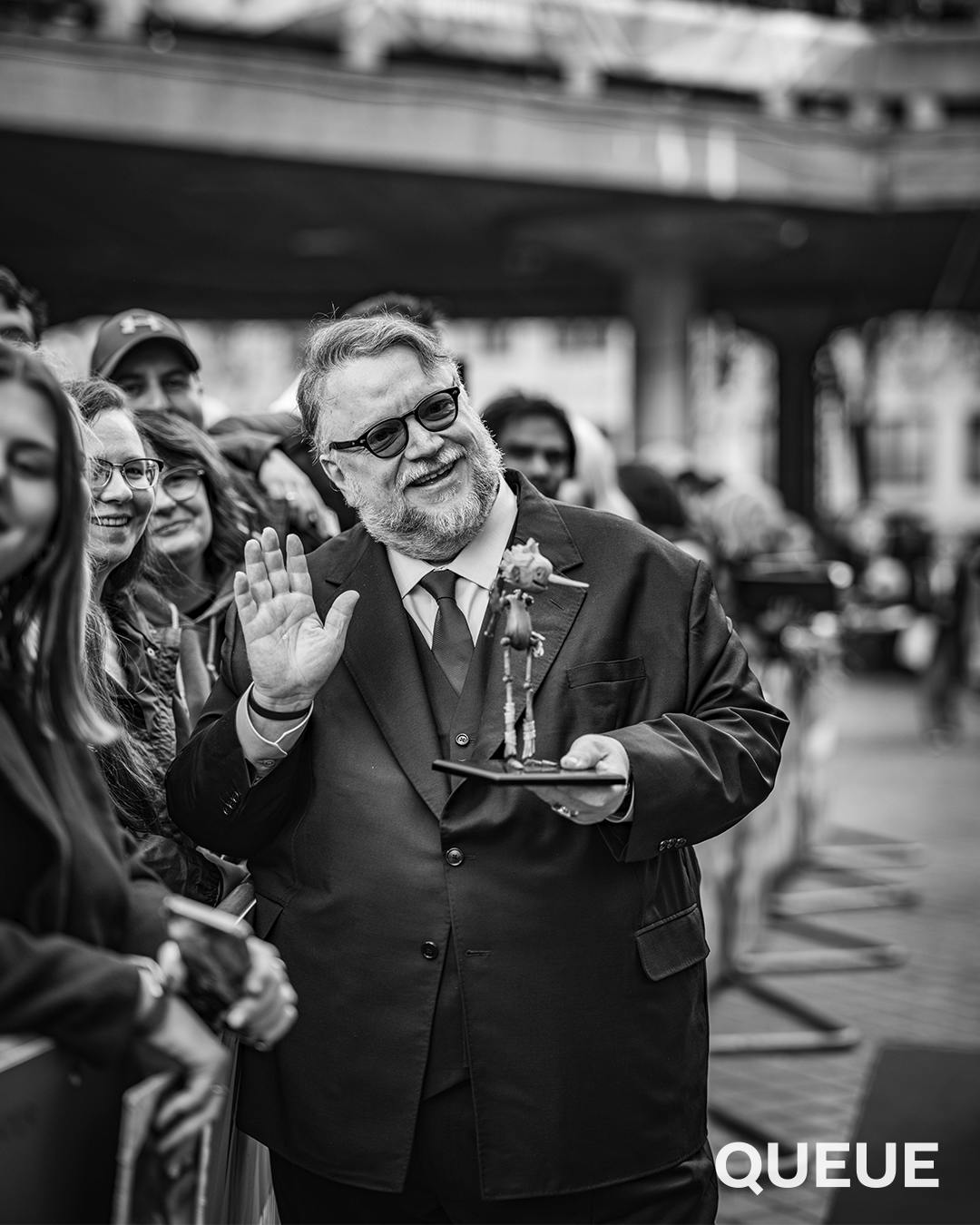 Guillermo del Toro wears a suit and waves from a crowd of people holding a miniature Pinocchio puppet.