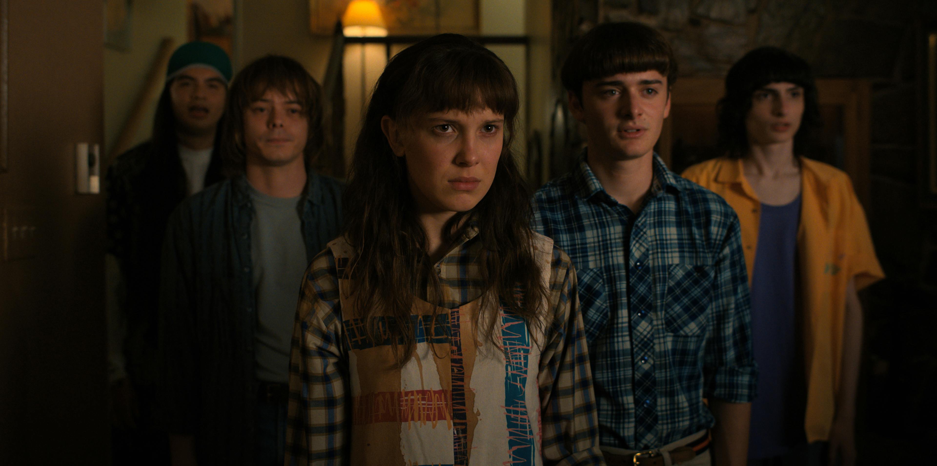 Eduardo Franco, Charlie Heaton, Millie Bobby Brown, Noah Schnapp, and Finn Wolfhard stand in a 'V' facing the camera. Franco wears a backwards blue hat, Heaton wears a grey t-shirt and flannel, Brown wears a flannel and a vest, Schnapp wears a flannel shirt, and Wolfhard wears an orange shirt over a navy tee. 