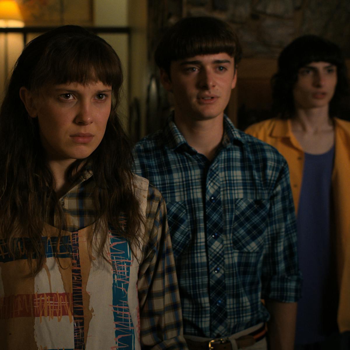 Eduardo Franco, Charlie Heaton, Millie Bobby Brown, Noah Schnapp, and Finn Wolfhard stand in a 'V' facing the camera. Franco wears a backwards blue hat, Heaton wears a grey t-shirt and flannel, Brown wears a flannel and a vest, Schnapp wears a flannel shirt, and Wolfhard wears an orange shirt over a navy tee. 