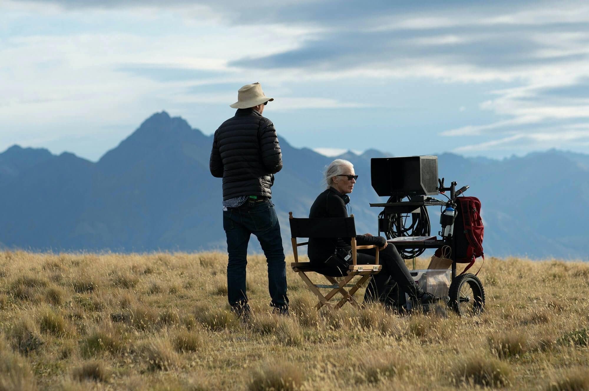 Producer Phil Jones and director Jane Campion stand in a windswept field, in dark clothing. In the background are dark blue mountains and a cloudy sky.