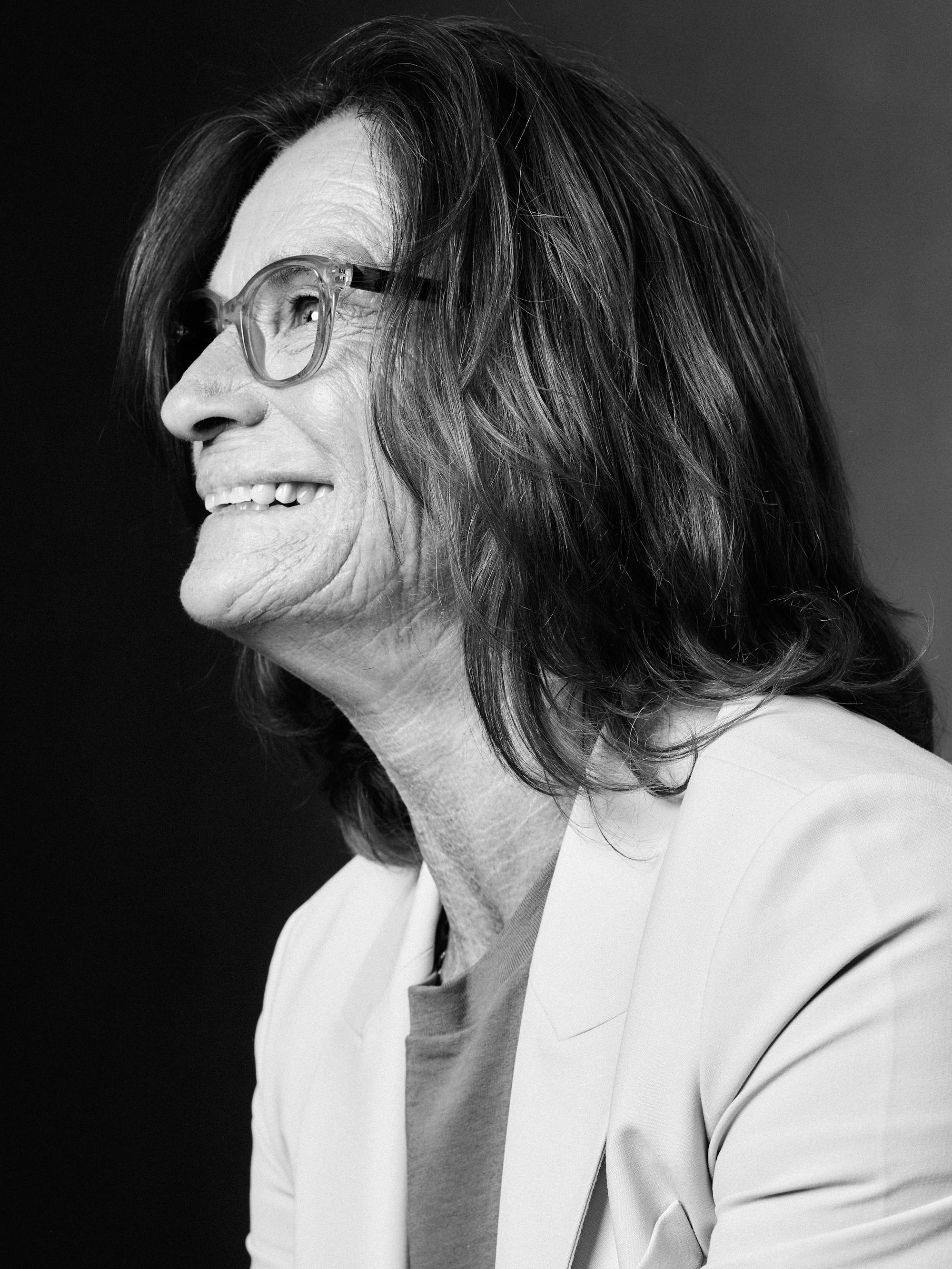 Bonnie Stoll wears a white blazer and glasses and smiles at something off-camera in the black-and-white image.