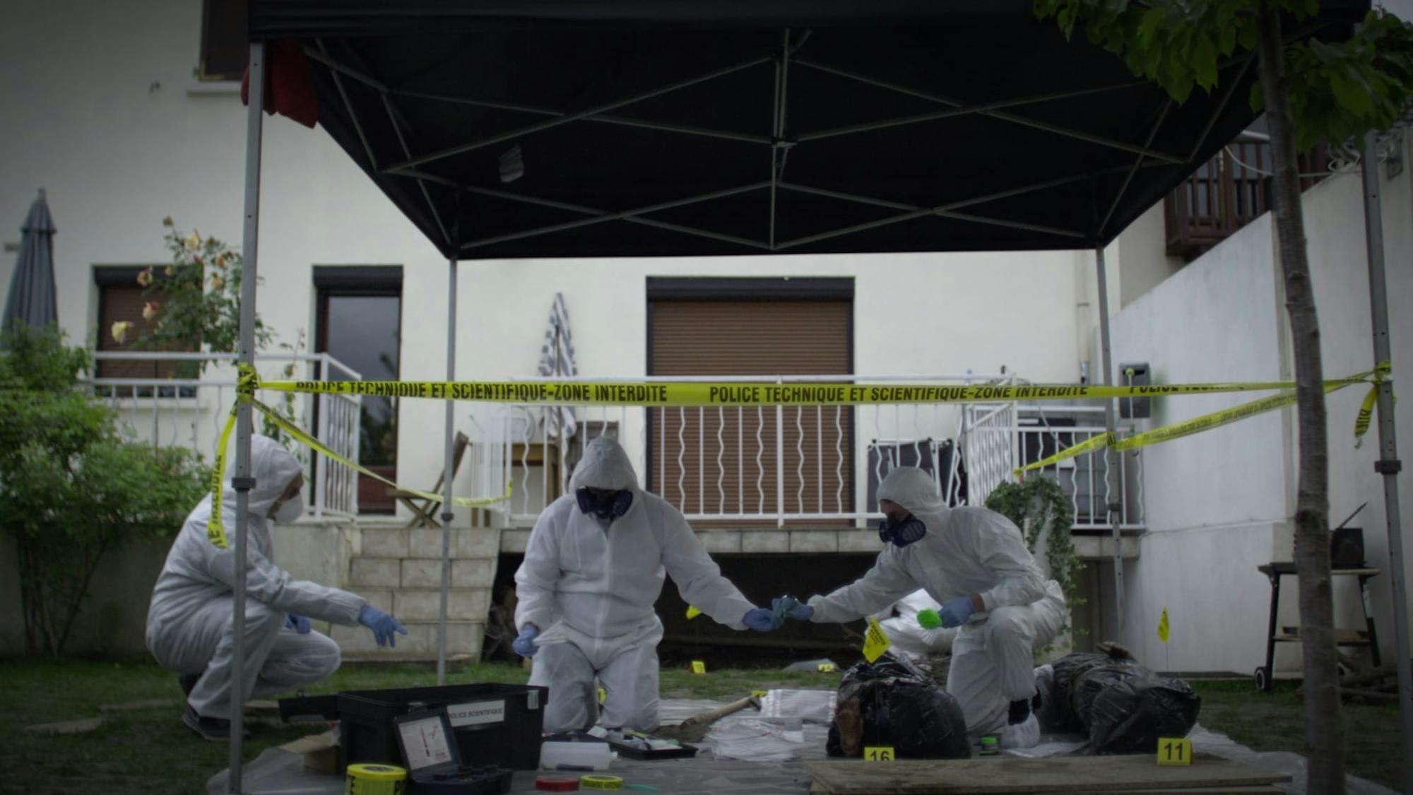 A crime scene. Three figures wear white hazmat suits and work within an area bounded with caution tape. The scene takes place outside of a white house and cement deck.