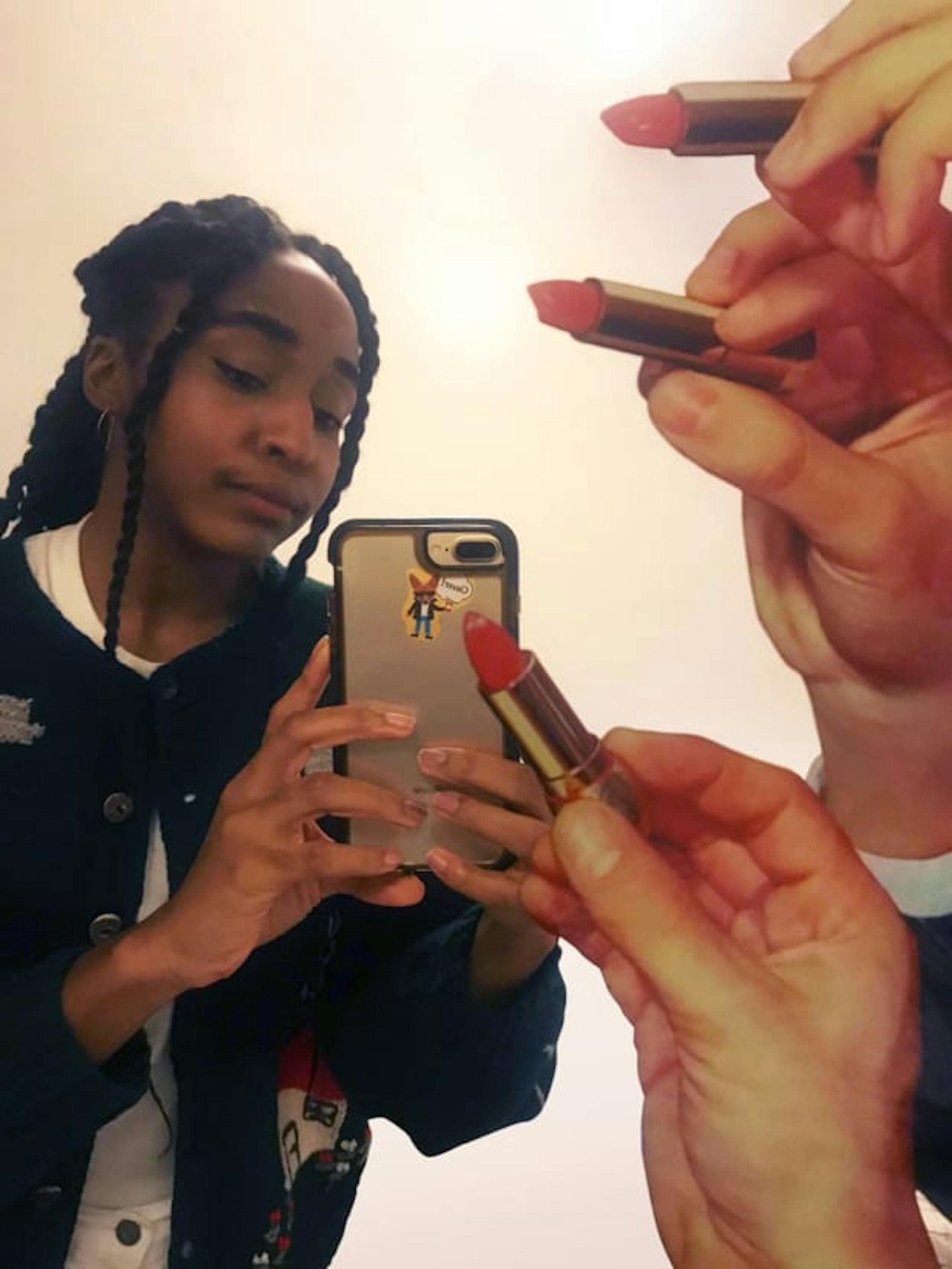 Voice actor Ayo Edebiri takes a mirror selfie as decals of hands holding lipstick tubes close in around her.