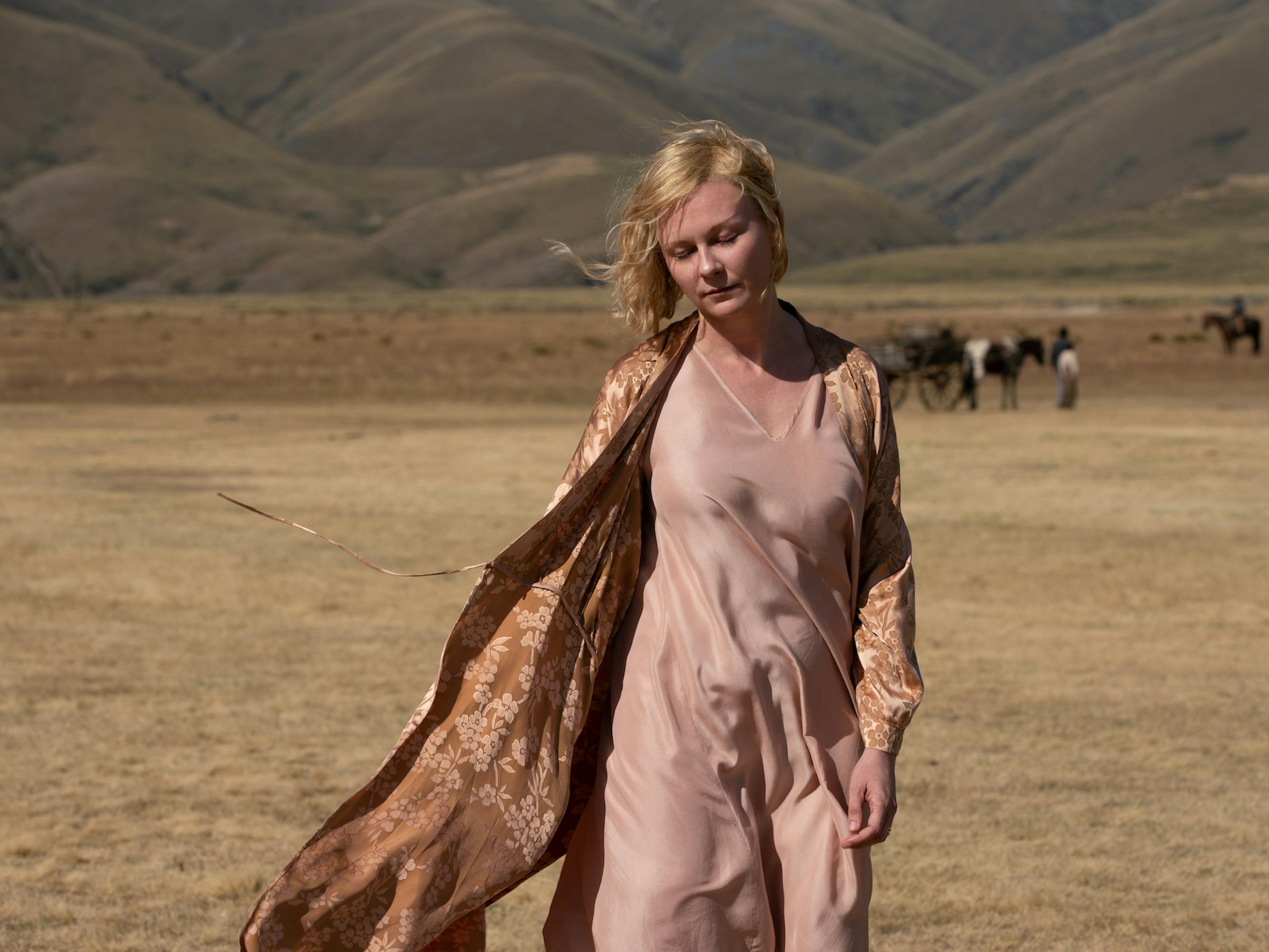 Rose (Kirsten Dunst) wears a long pink dress and brown floral robe as she stands in a dusty expanse with some horses behind her.