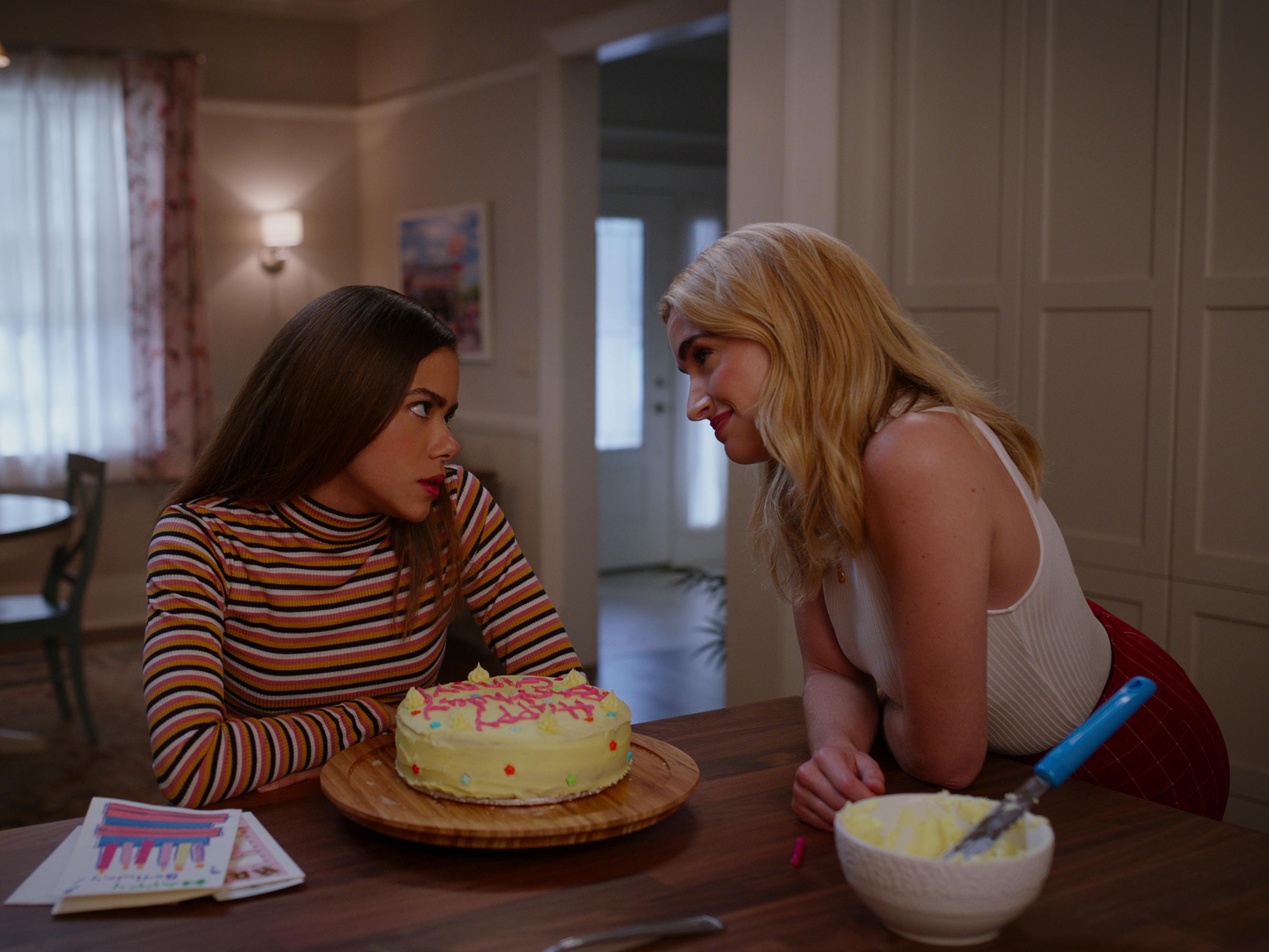 Ginny and Georgia sit at a kitchen counter with a birthday cake between them. Ginny wears a striped turtleneck, and Georgia wears a white tank. 