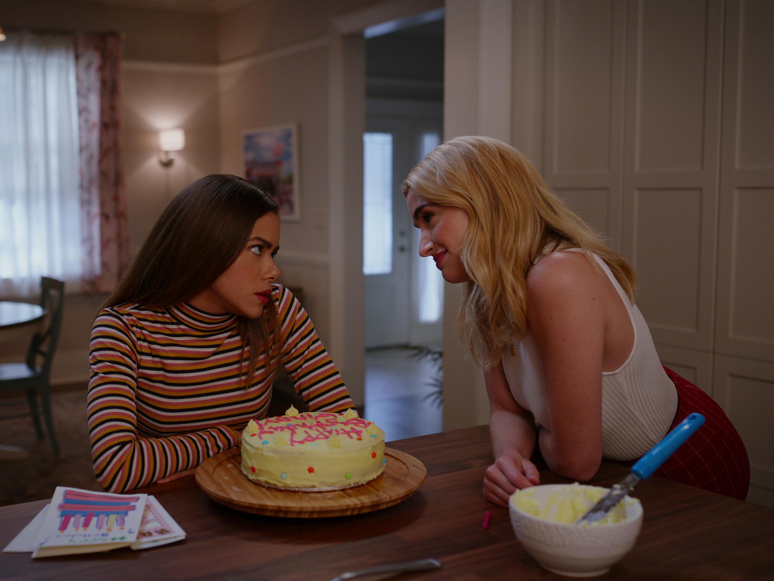 Ginny and Georgia sit at a kitchen counter with a birthday cake between them. Ginny wears a striped turtleneck, and Georgia wears a white tank.