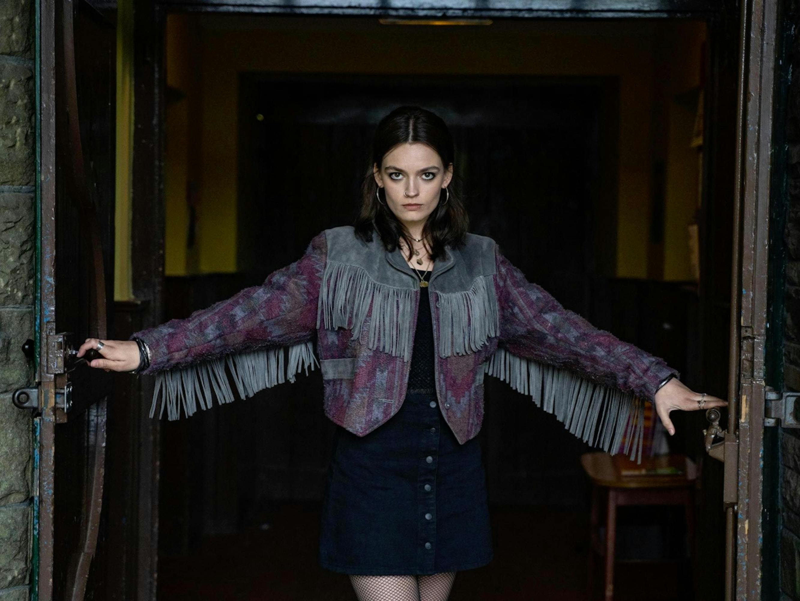 Maeve Wiley (Emma Mackey) wears a dark skirt, fringe coat, and tights as she opens the doors with an ominous expression on her face.