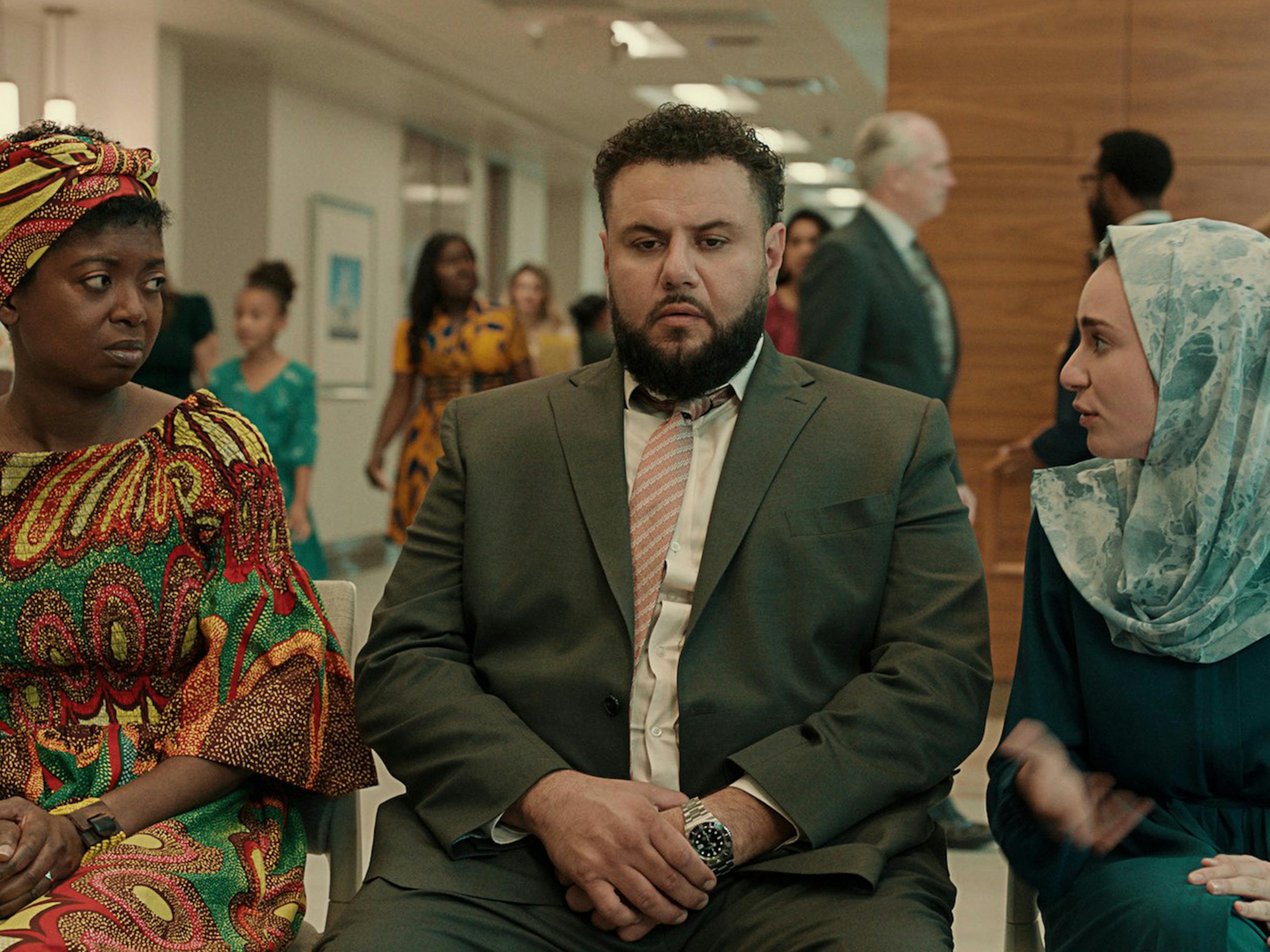 Aisha (Nene Nwoko), Mo (Mo Amer), and Maysoon (Zein Khleif) sit together. Aisha wears a matching scarf and dress, Mo wears a suit, and Maysoon wears a teal top and patterned scarf.