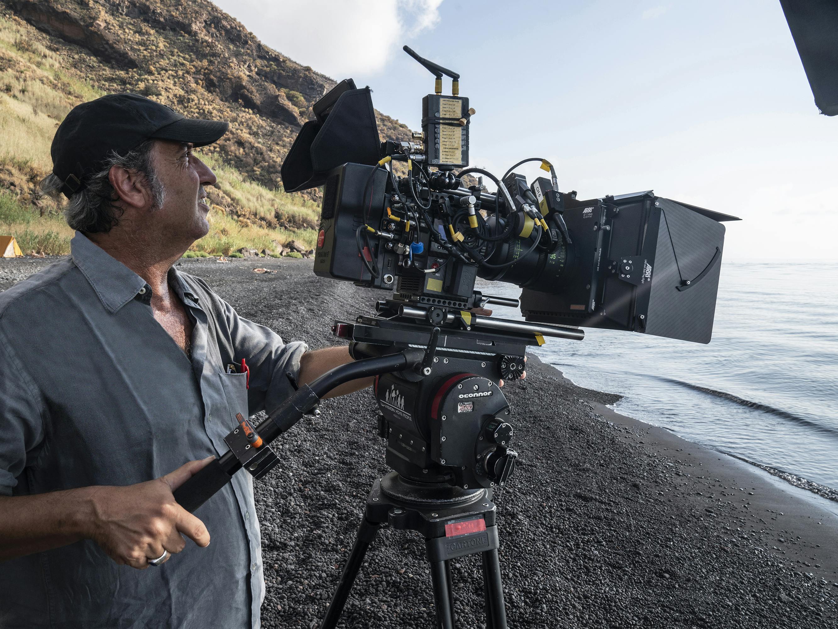Paolo Sorrentino works a camera on a beach in a grey shirt and black hat.