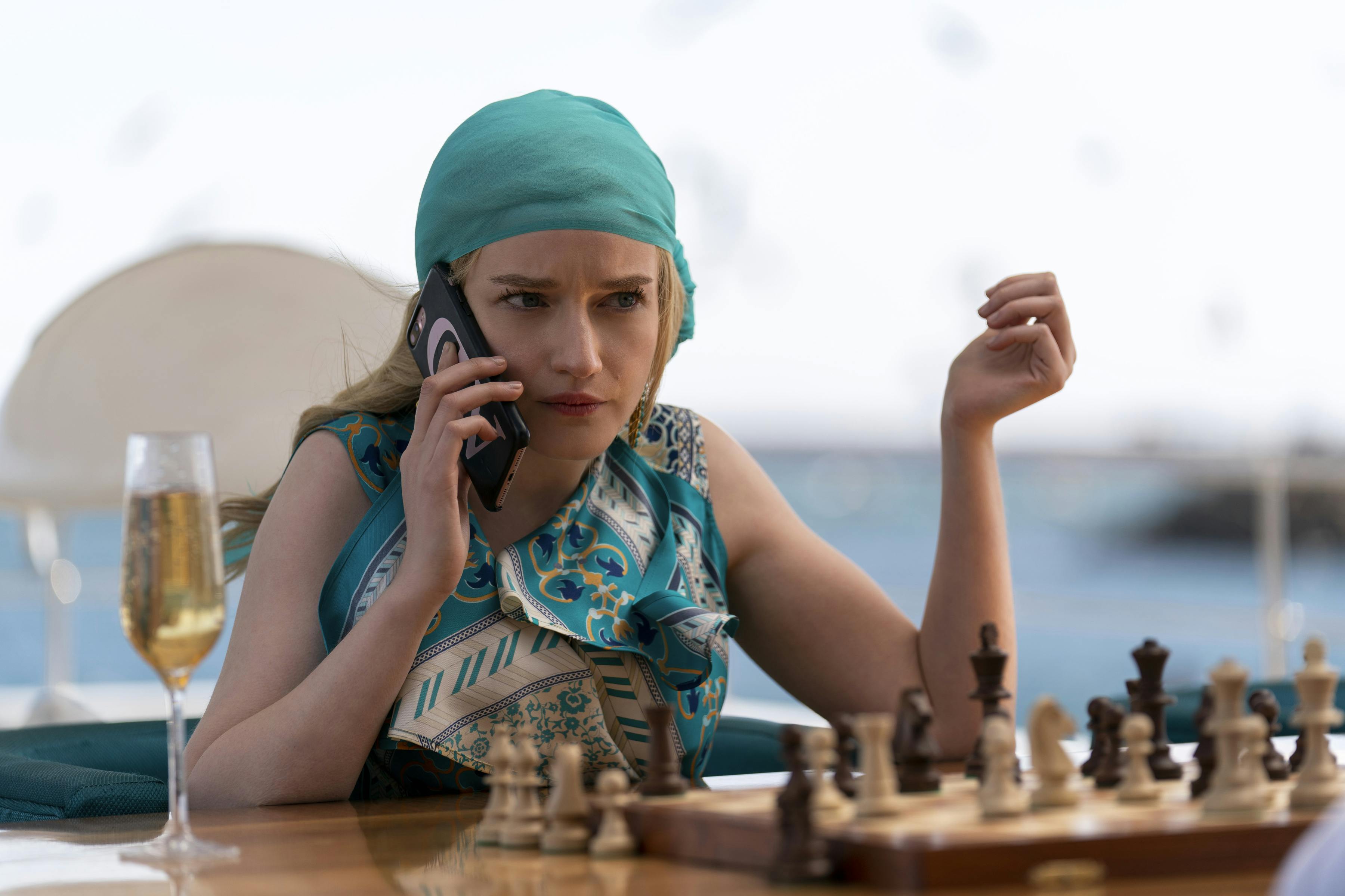 Anna Delvey, played by Julia Garner, wears a turquoise headscarf and patterned shirt. In front of her sits a chess board and a glass of champagne.
