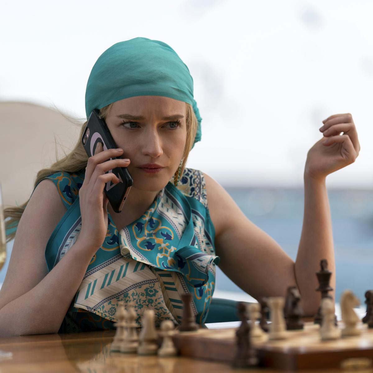 Anna Delvey, played by Julia Garner, wears a turquoise headscarf and patterned shirt. In front of her sits a chess board and a glass of champagne.