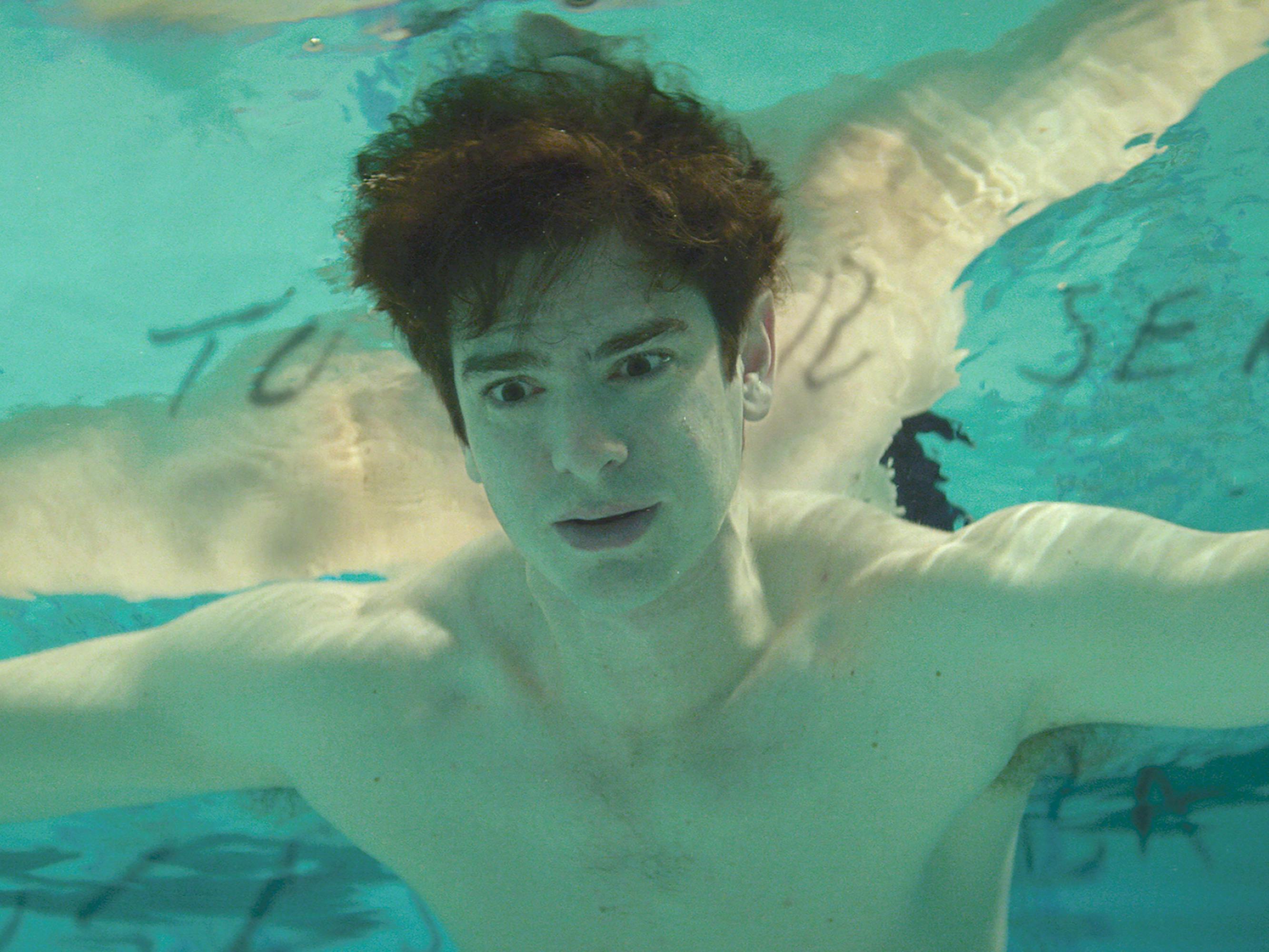 Andrew Garfield floats in a pool looking existential. Floating letters surround his face. He looks scary with his eyes open under water.
