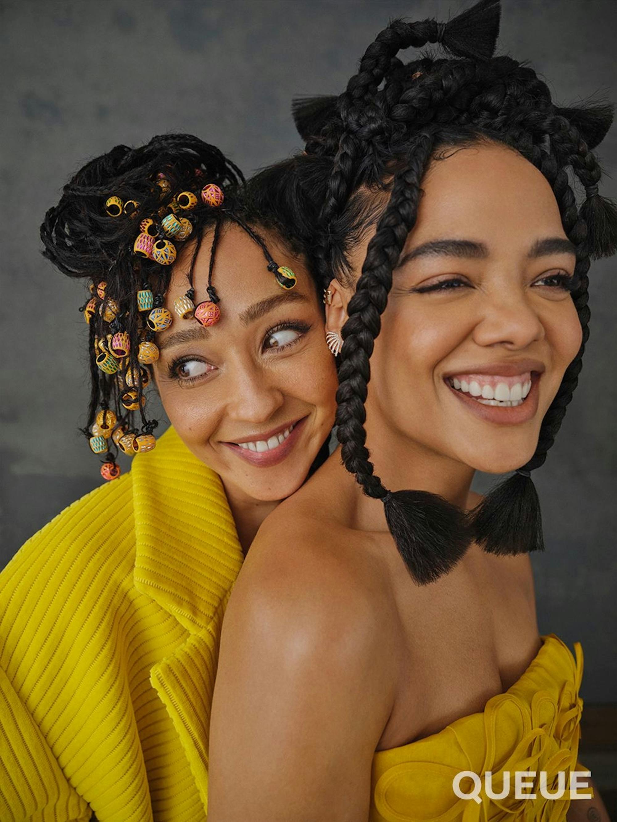 Ruth Negga wears a yellow coat and smiles as she stands behind Tessa Thomspon. Thompson also wears a yellow top and has a big grin.