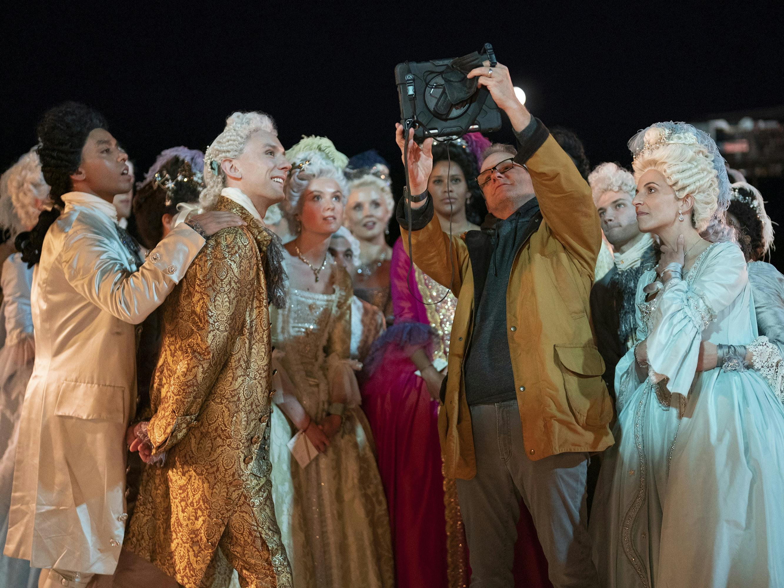 Tom Verica behind the scenes of Episode 6. He holds a monitor to a crowd of actors all dressed in fabulous costumes.