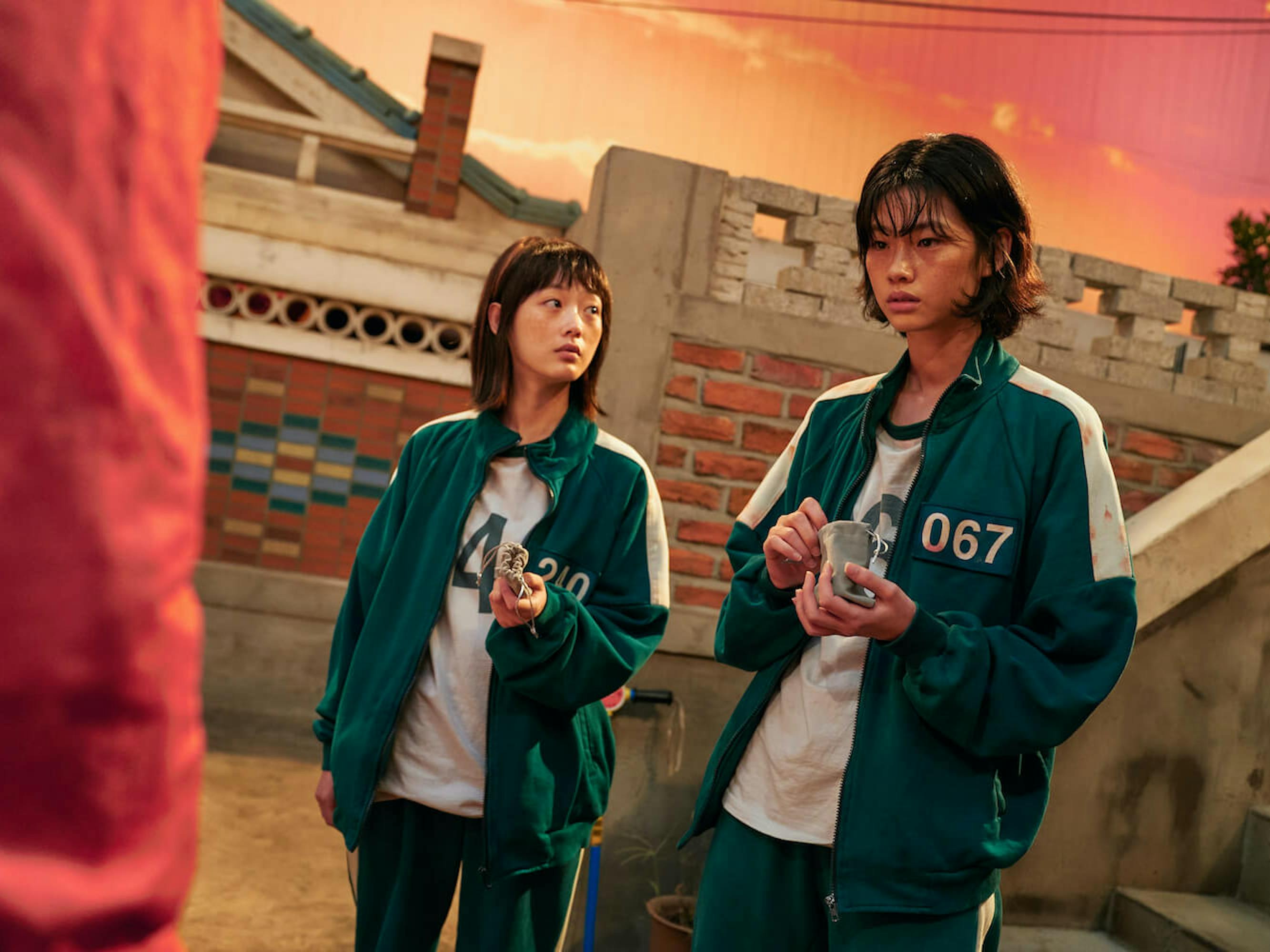 Ji-yeong (Lee Yoo-mi) and Kang Sae-byeok (Jung Ho-yeon) stand together picking something out of a small leather bag. Their green tracksuits are unzipped and they are sweaty and afraid. In the background is a brick wall and a red and orange sky.