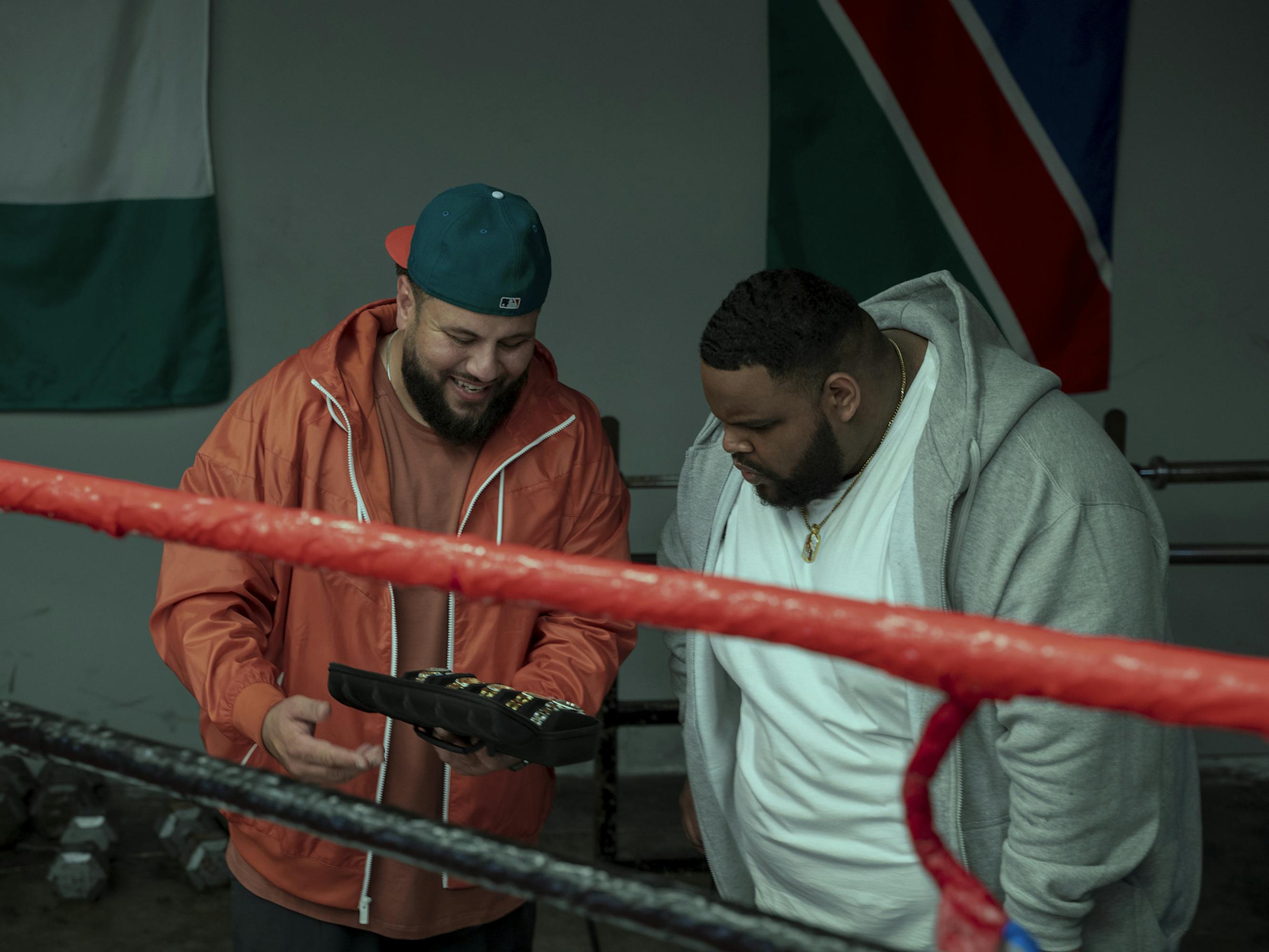 Mo (Mo Amer) and Dante (Rafael Castillo) look at watches by a boxing ring, with flags hanging on the walls.